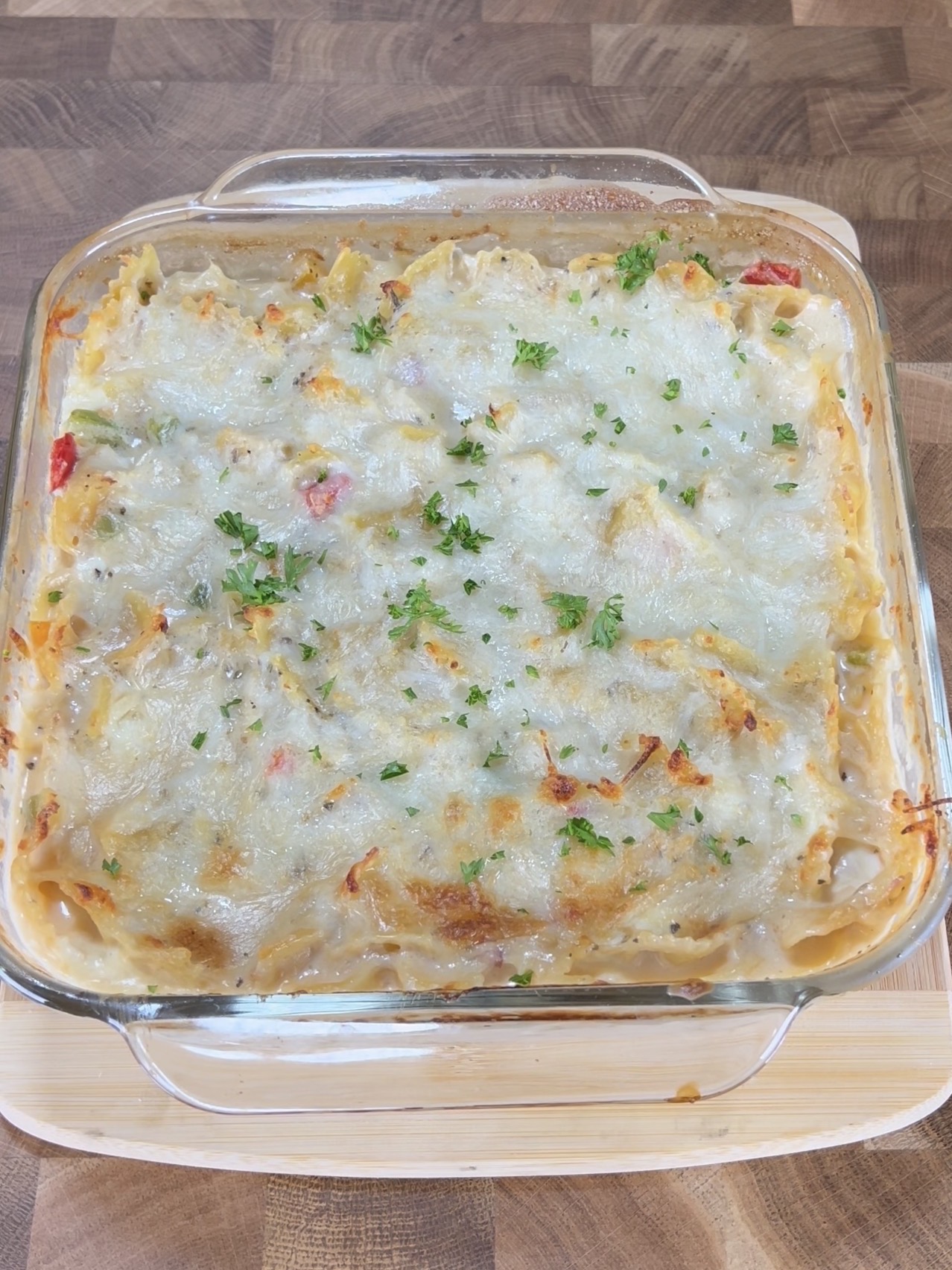 A glass baking dish filled with a baked casserole topped with melted, slightly browned cheese and garnished with chopped parsley, sitting on a wooden cutting board.