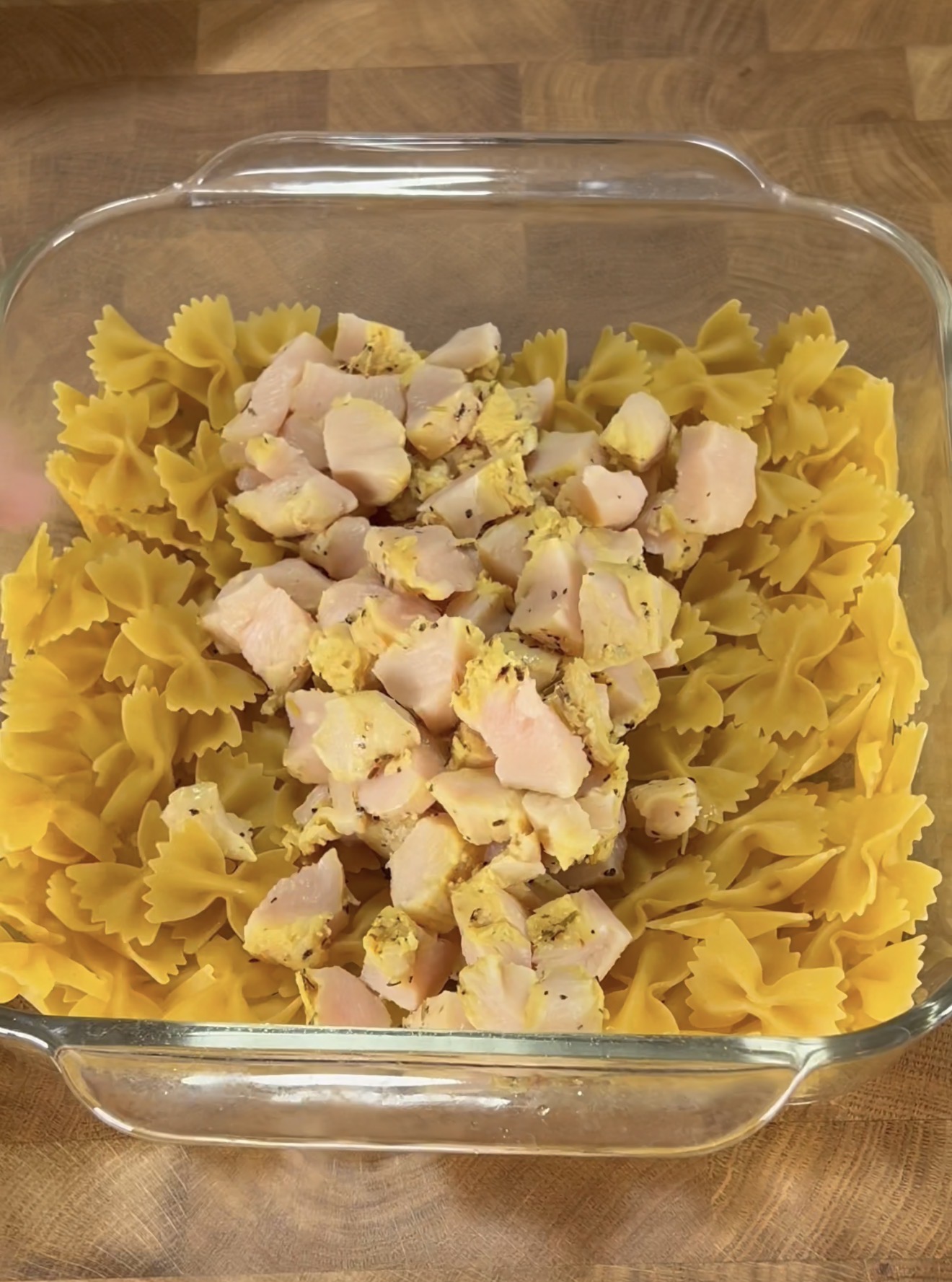 A glass baking dish on a wooden surface holds uncooked farfalle pasta topped with cubes of cooked, seasoned chicken.