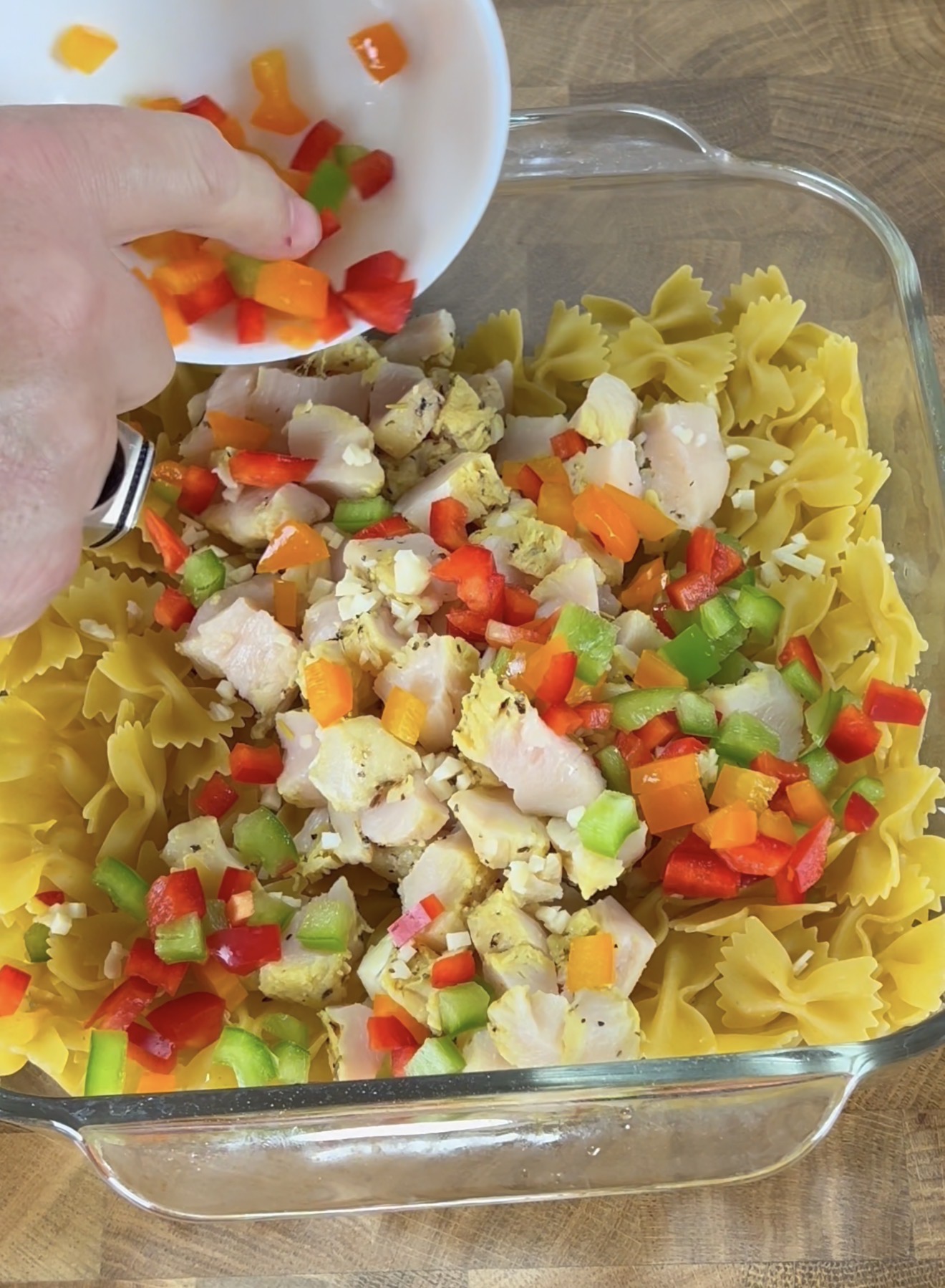 A hand sprinkles chopped red, orange, and green bell peppers from a white bowl over cooked bowtie pasta and diced chicken in a glass baking dish on a wooden surface.