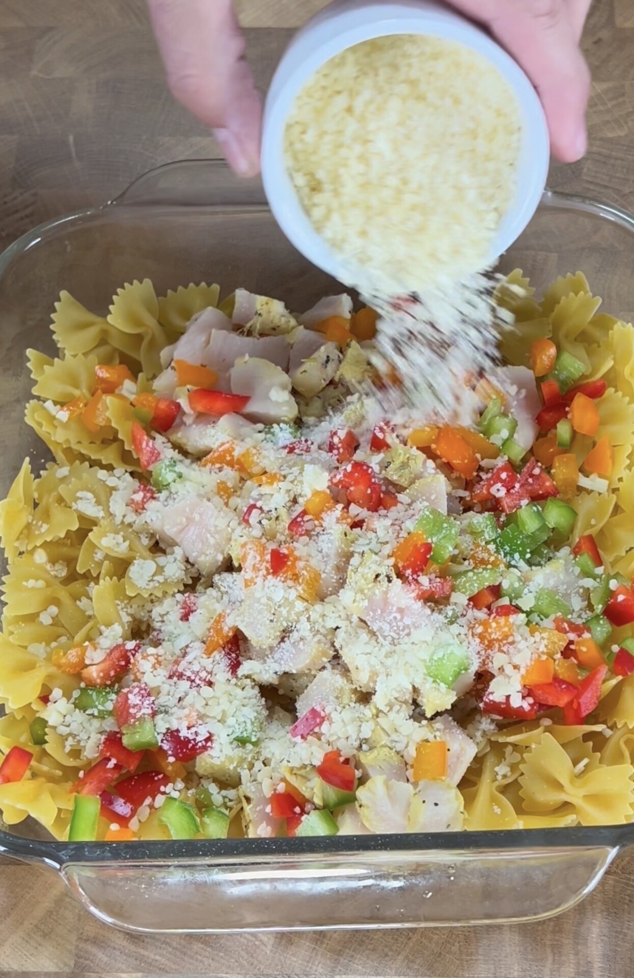 A hand sprinkles grated cheese from a small bowl over uncooked bowtie pasta, diced chicken, and chopped red and green bell peppers in a glass baking dish.