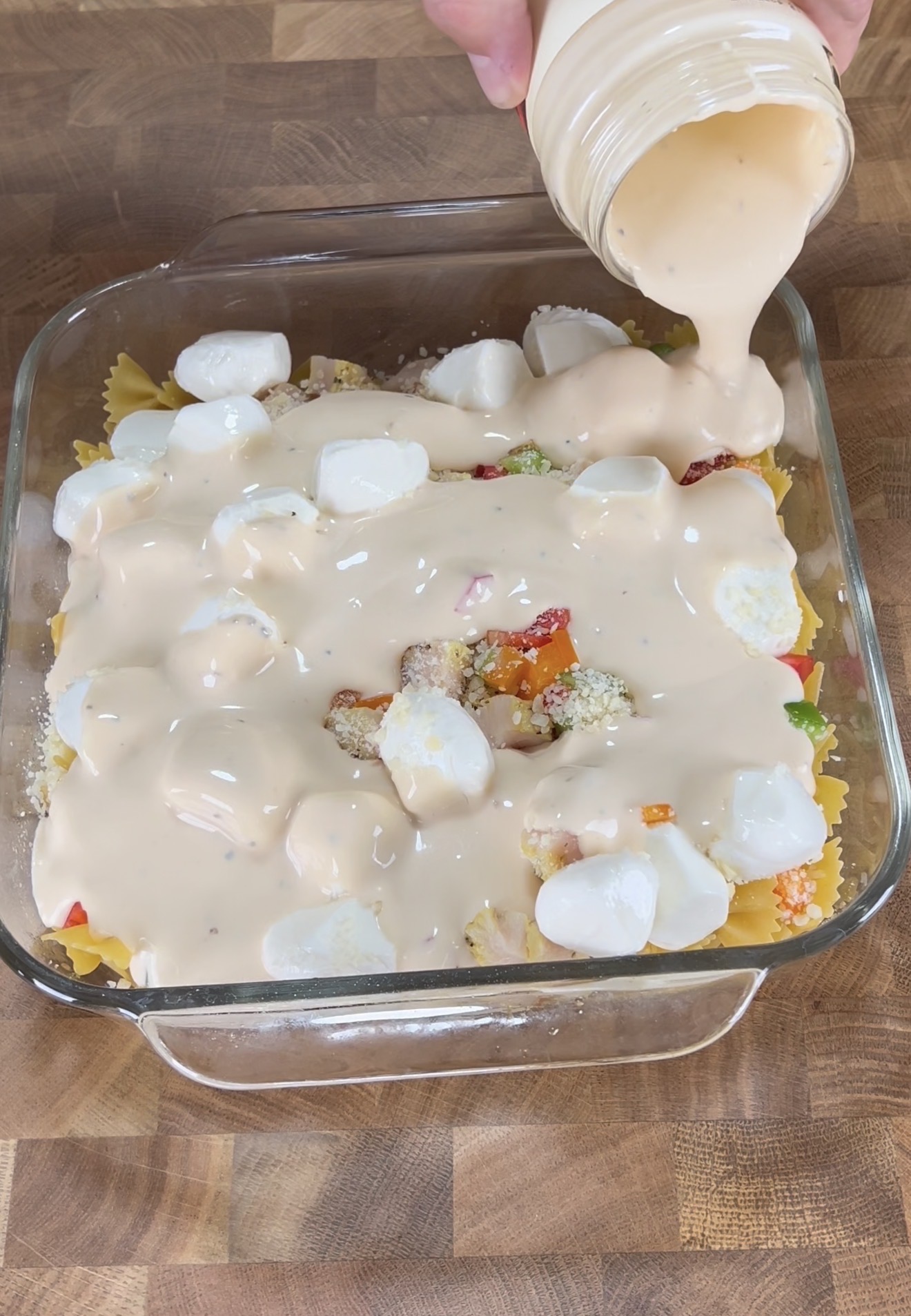 A hand pours creamy dressing from a jar over a glass baking dish filled with uncooked pasta, vegetables, croutons, and mozzarella balls on a wooden surface.