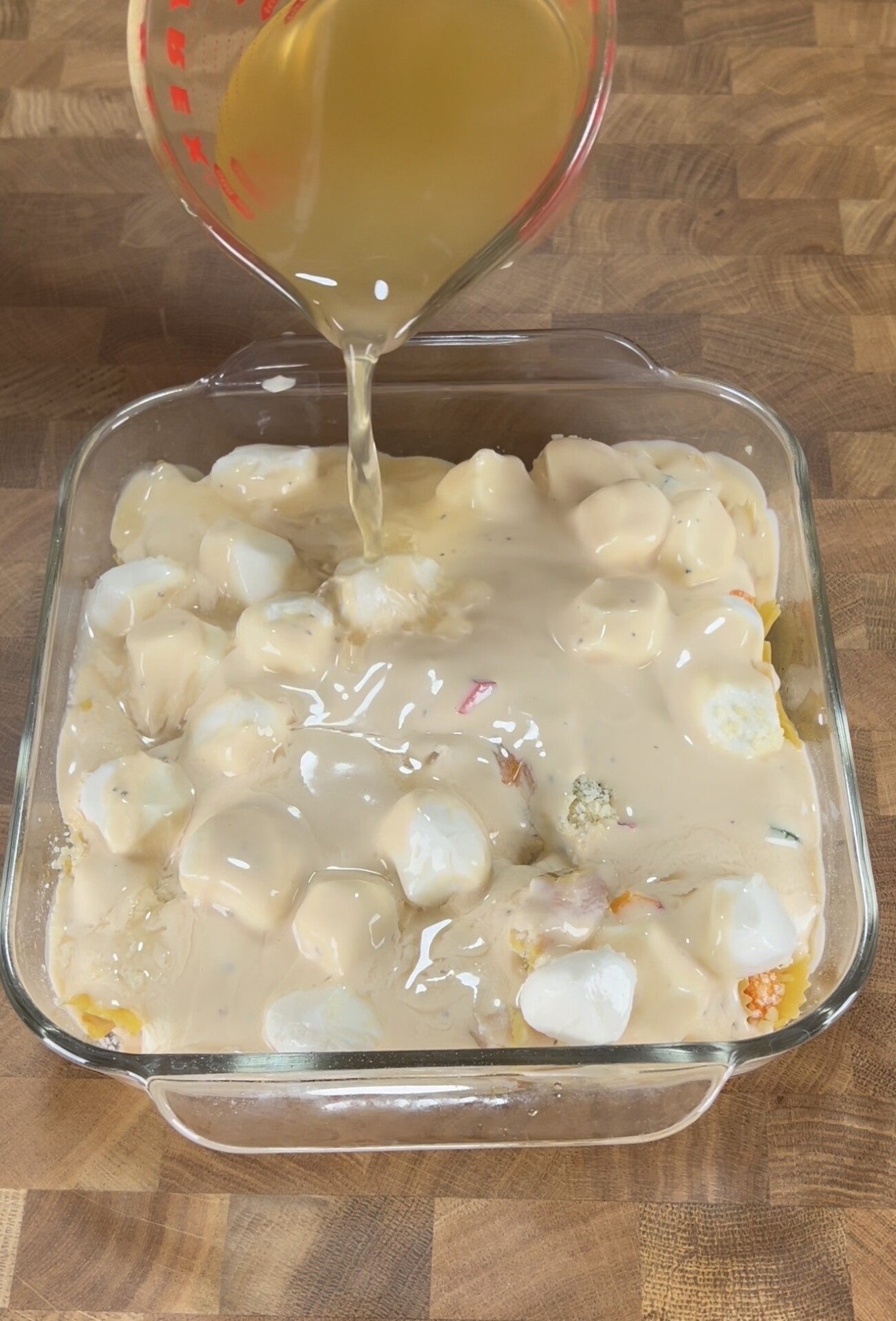 A glass measuring cup pours a light brown liquid over a baking dish filled with white sauce, chunks, and colorful bits, sitting on a wooden surface.