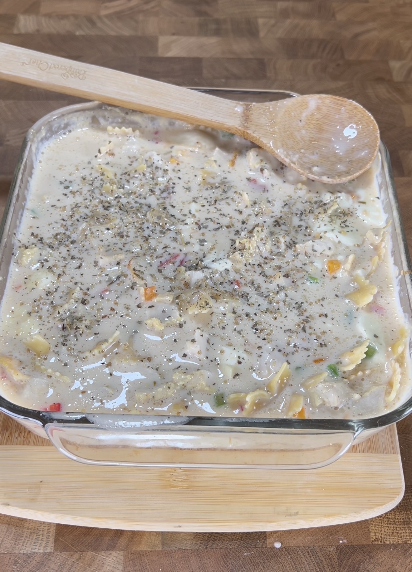 A glass baking dish filled with an uncooked creamy pasta casserole topped with dried herbs. A wooden spoon rests on the dish, and everything sits on a wooden surface.