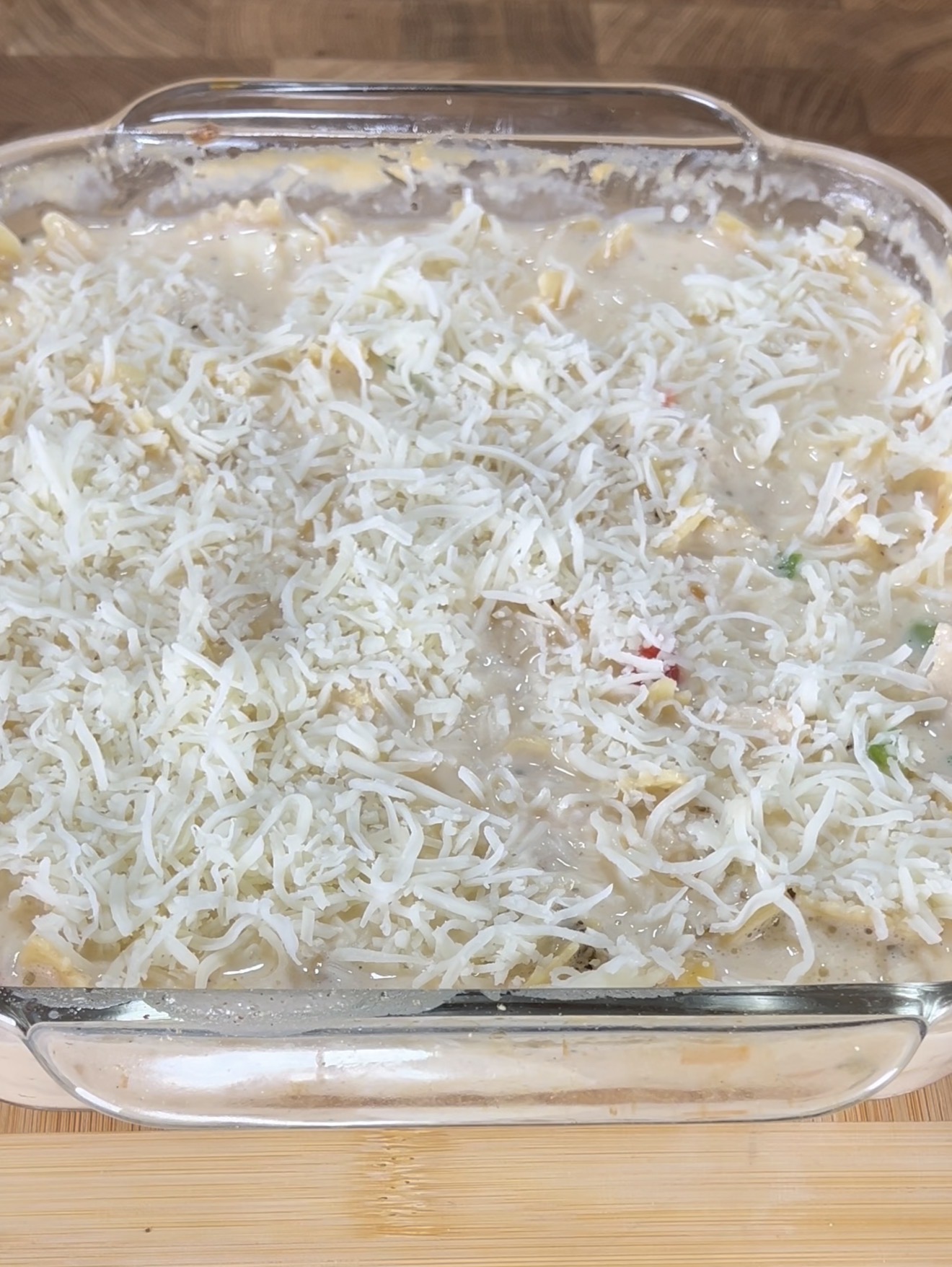 A glass baking dish filled with an unbaked casserole topped with shredded cheese, sitting on a wooden surface.