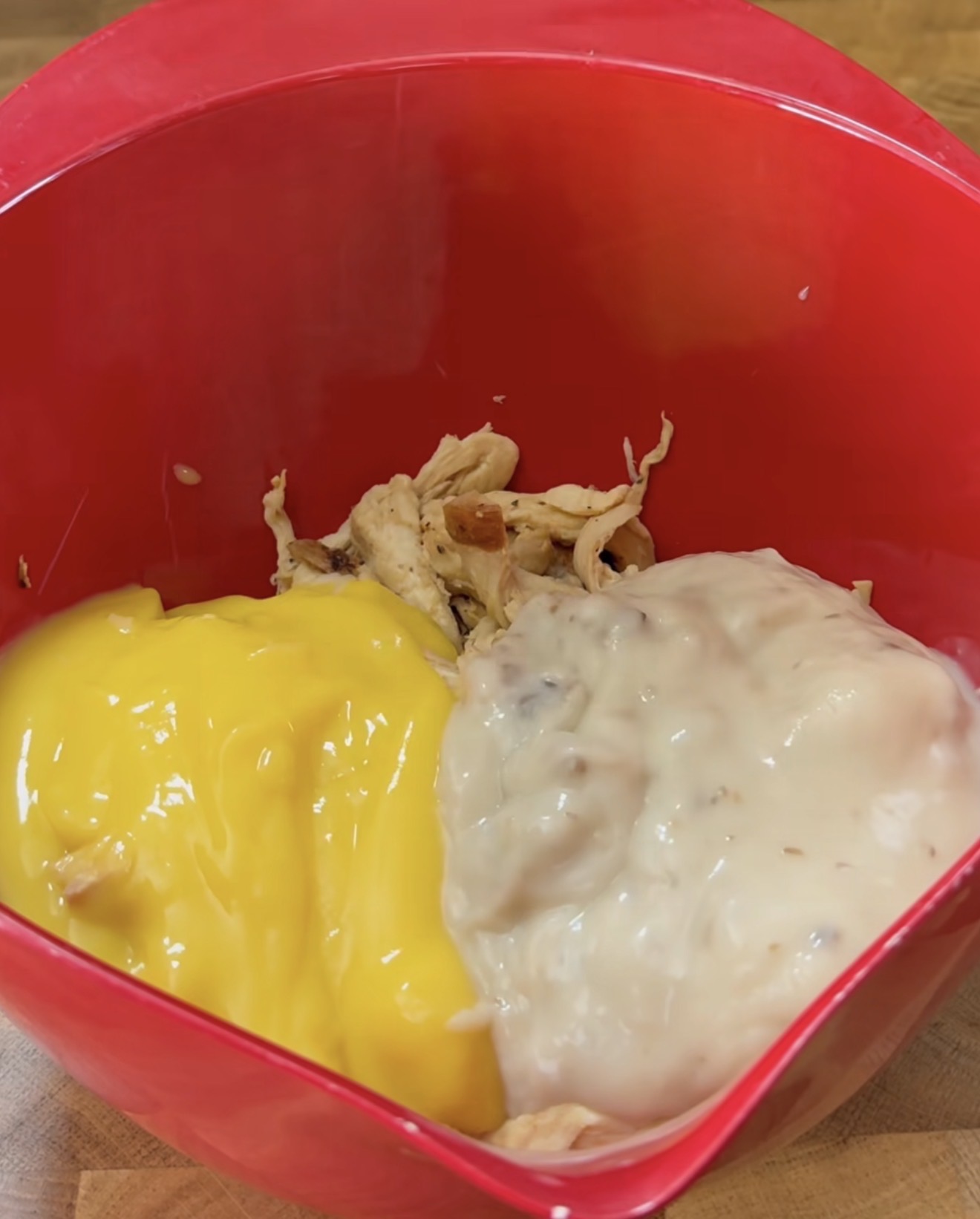 A red mixing bowl containing shredded chicken, a portion of yellow soup or sauce, and a portion of white creamy soup or sauce, on a wooden surface.