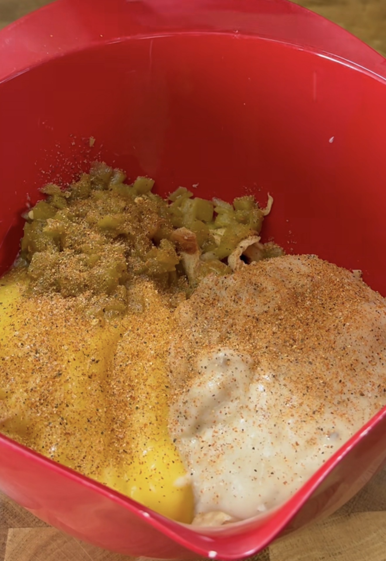 A red mixing bowl containing shredded chicken, chopped green chilies, canned cream soup, shredded cheese, and a sprinkling of seasoning.