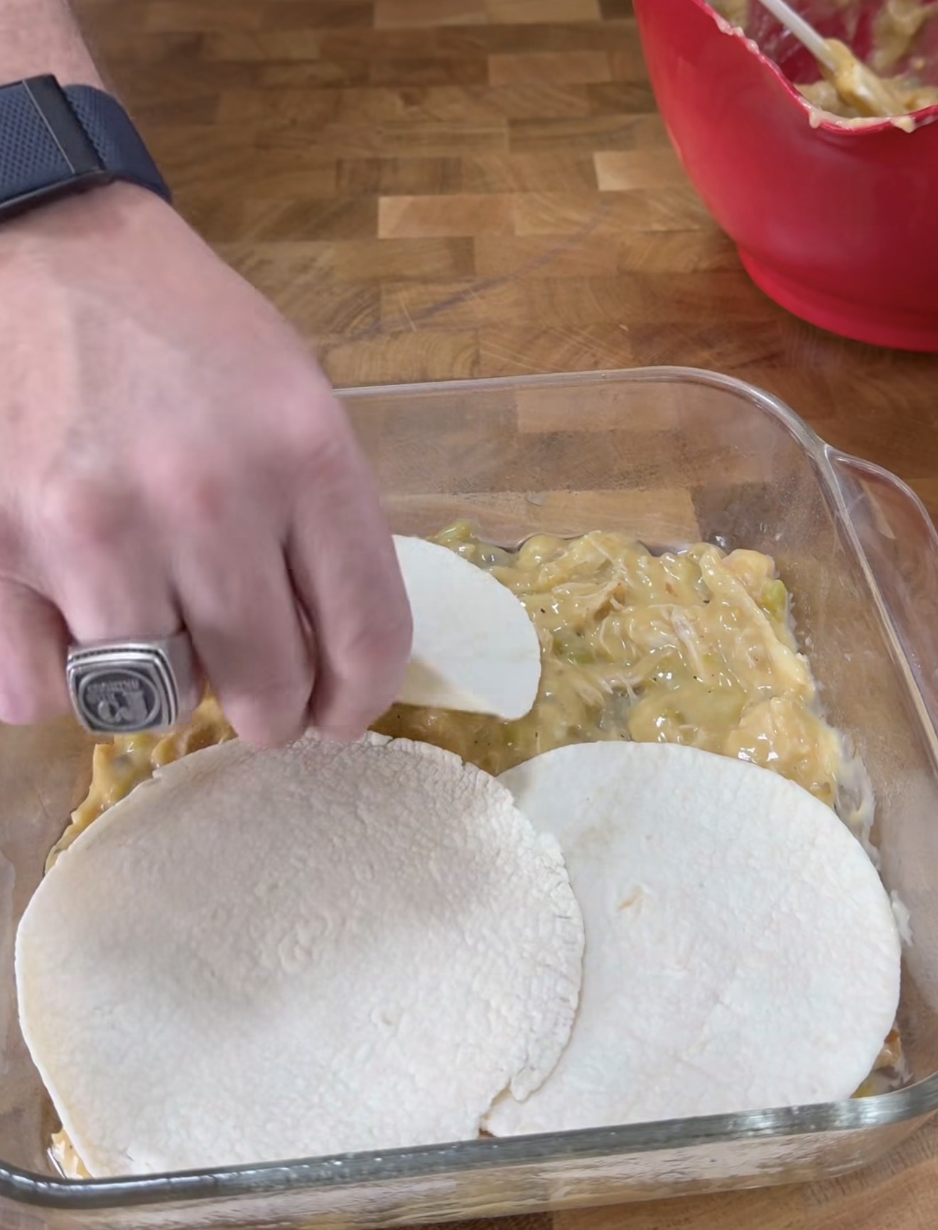 A hand places white corn tortillas over a layer of creamy filling in a glass baking dish, with a red mixing bowl in the background on a wooden surface.