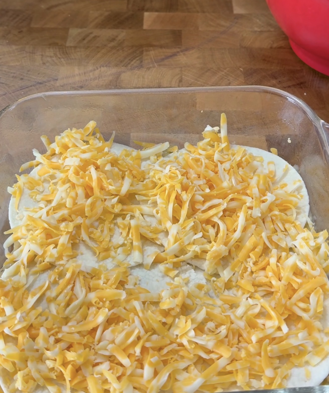 A glass baking dish filled with an even layer of shredded yellow and white cheese on top of a creamy mixture, set on a wooden countertop. A red mixing bowl is partially visible in the background.