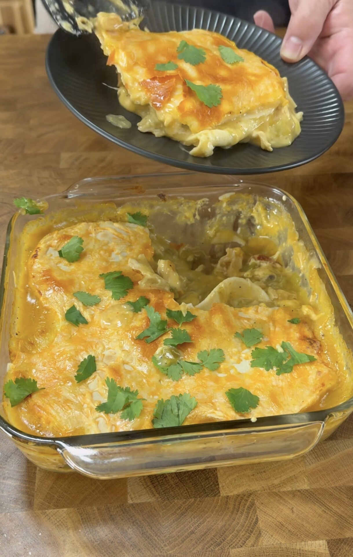 A glass baking dish filled with cheesy lasagna topped with fresh cilantro. A hand is serving a slice onto a black plate over a wooden surface.