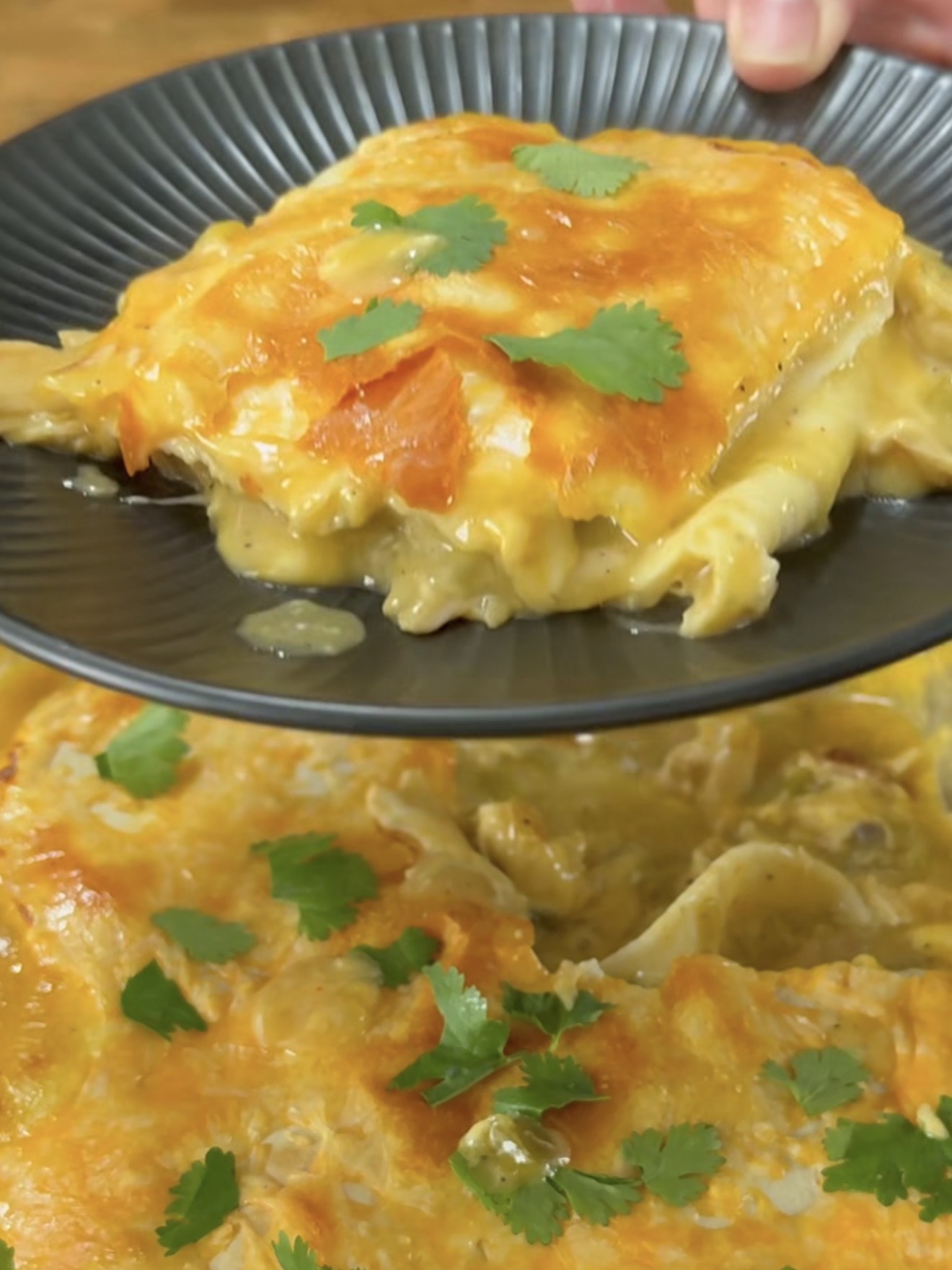 A serving of cheesy baked enchiladas topped with fresh cilantro is being lifted onto a black plate, with the rest of the enchilada dish visible below.
