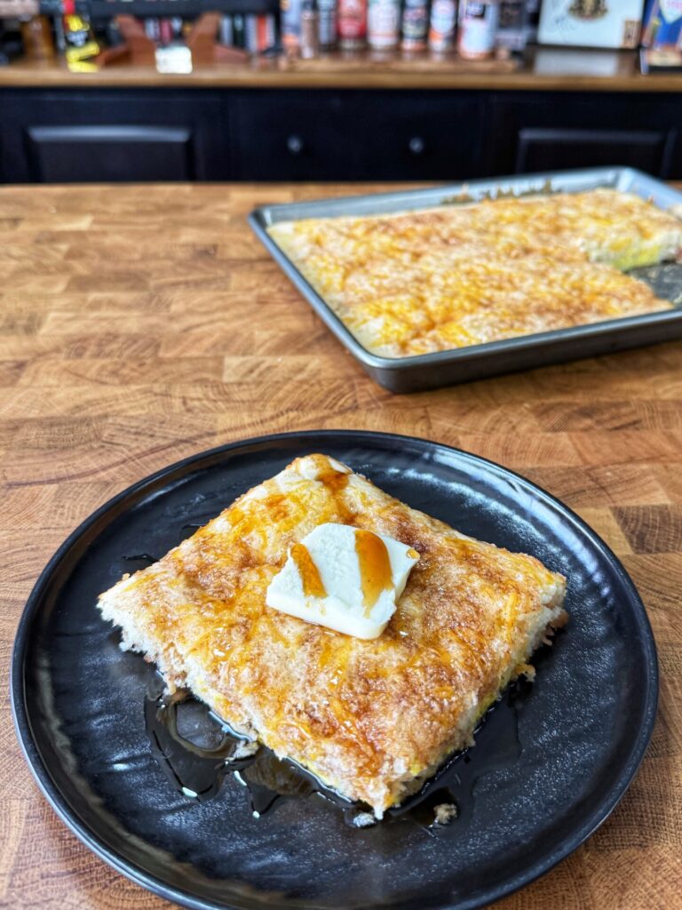A square piece of golden, flaky pastry topped with a pat of butter and drizzled with syrup sits on a black plate; a tray with more pastry is visible on a wooden countertop in the background.
