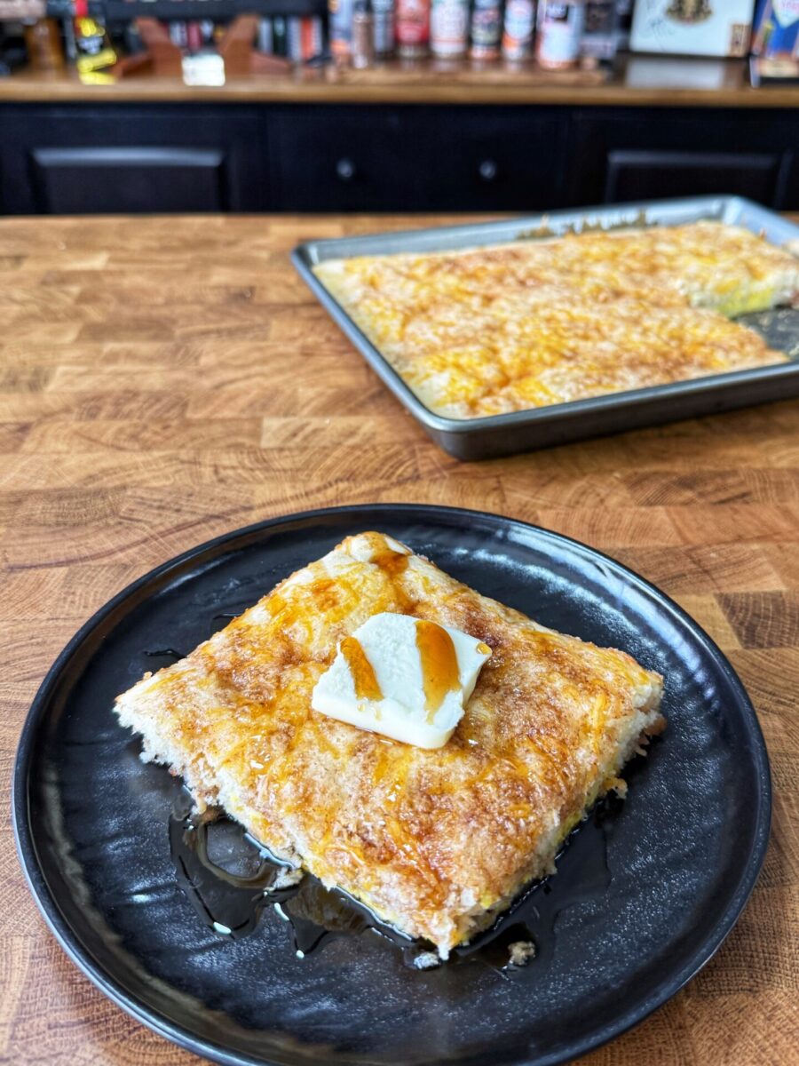 A square piece of golden, flaky pastry topped with a pat of butter and drizzled with syrup sits on a black plate; a tray with more pastry is visible on a wooden countertop in the background.