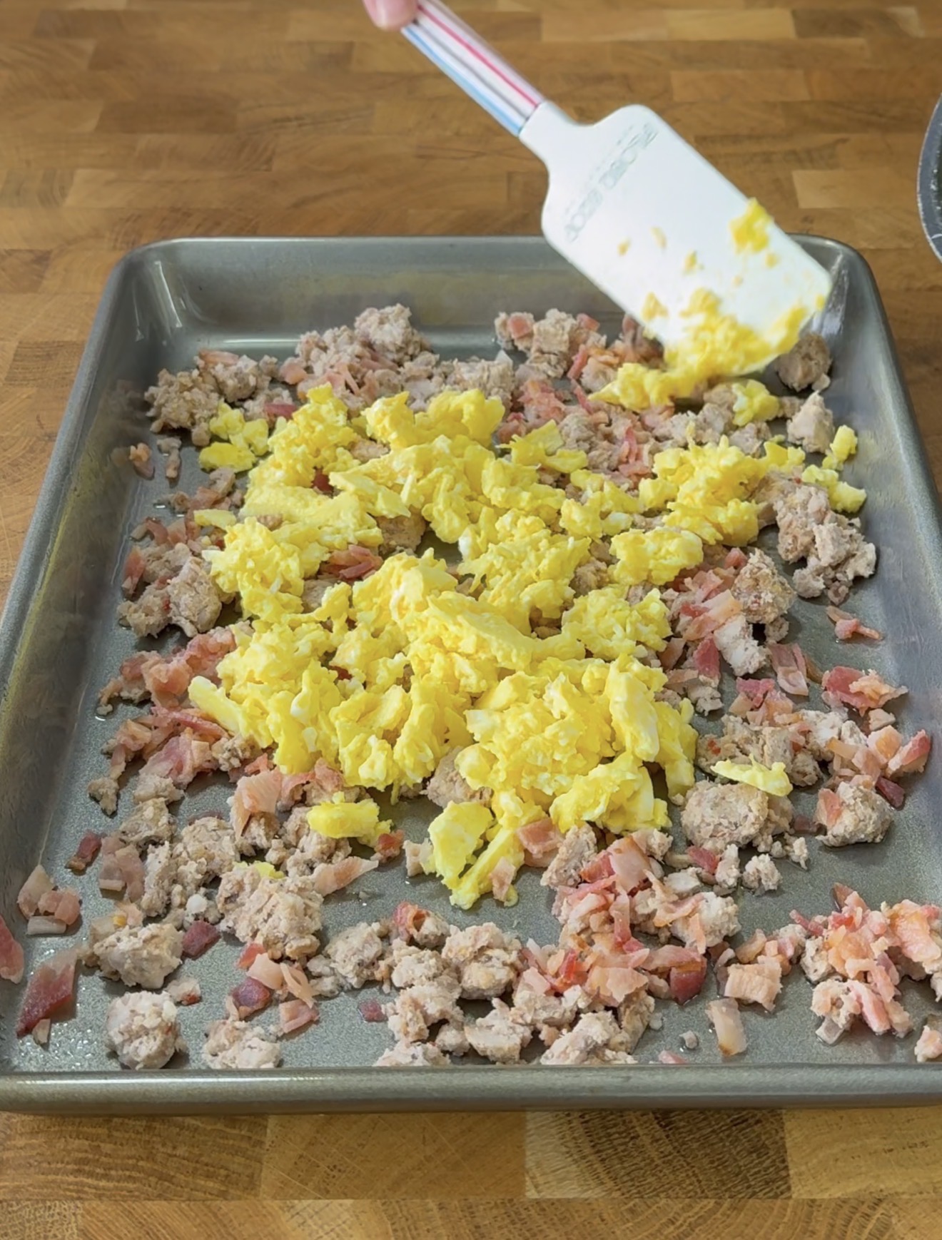 A baking sheet on a wooden surface holds cooked scrambled eggs, crumbled sausage, and chopped bacon. A hand using a white spatula is stirring or spreading the ingredients.