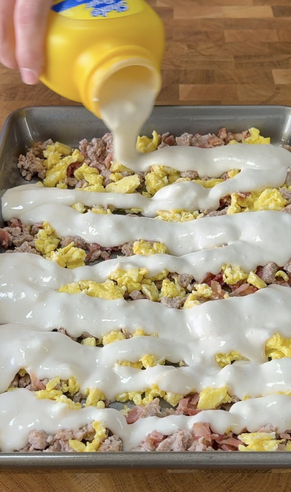 A hand pours creamy white sauce from a yellow bottle over a baking tray filled with scrambled eggs, cooked sausage, and bacon on a wooden counter.