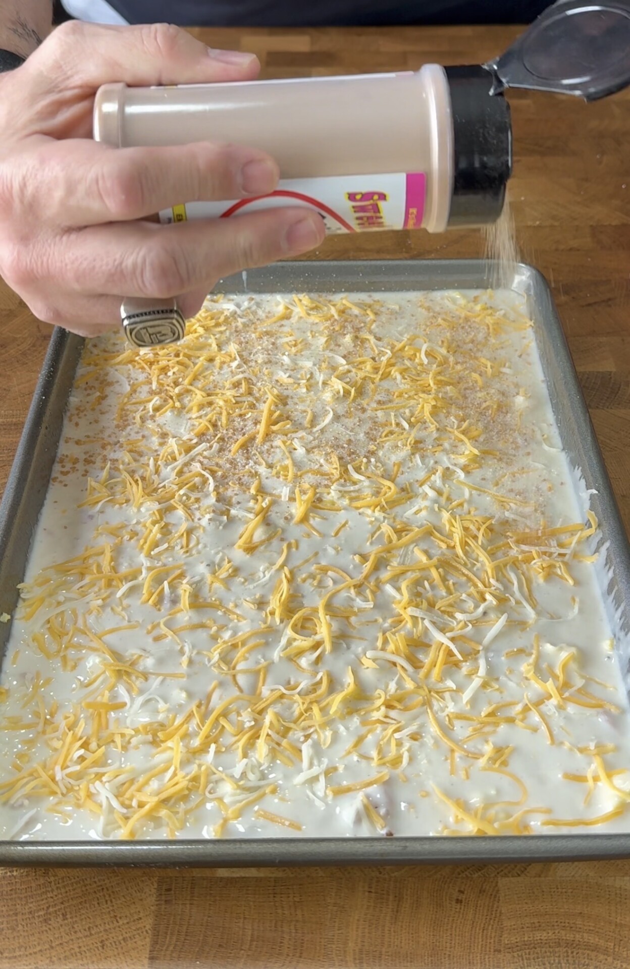 A hand sprinkles seasoning from a shaker over a baking tray filled with a creamy mixture topped with shredded cheese, resting on a wooden surface.