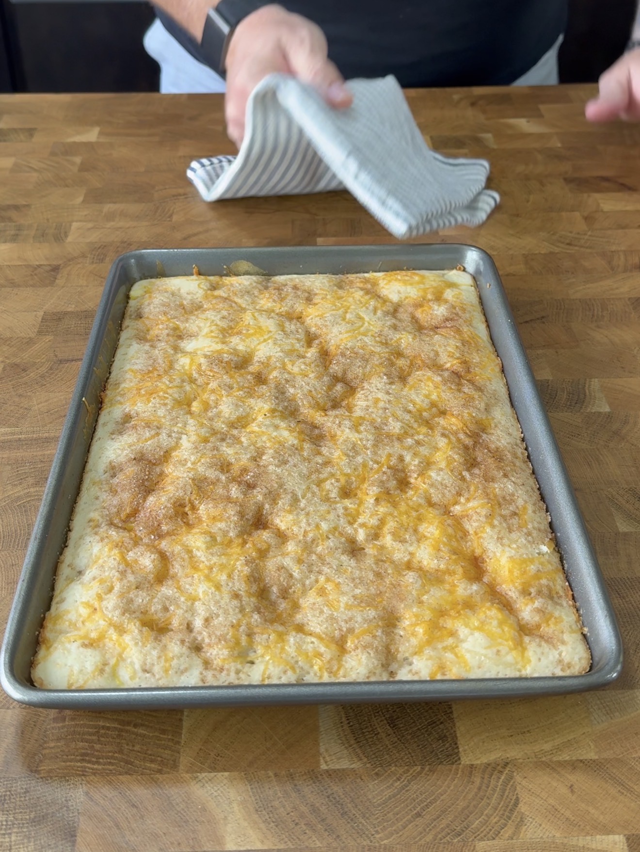 A person holds a striped towel above a baking sheet with a freshly baked, golden-brown cheesy bread on a wooden countertop.
