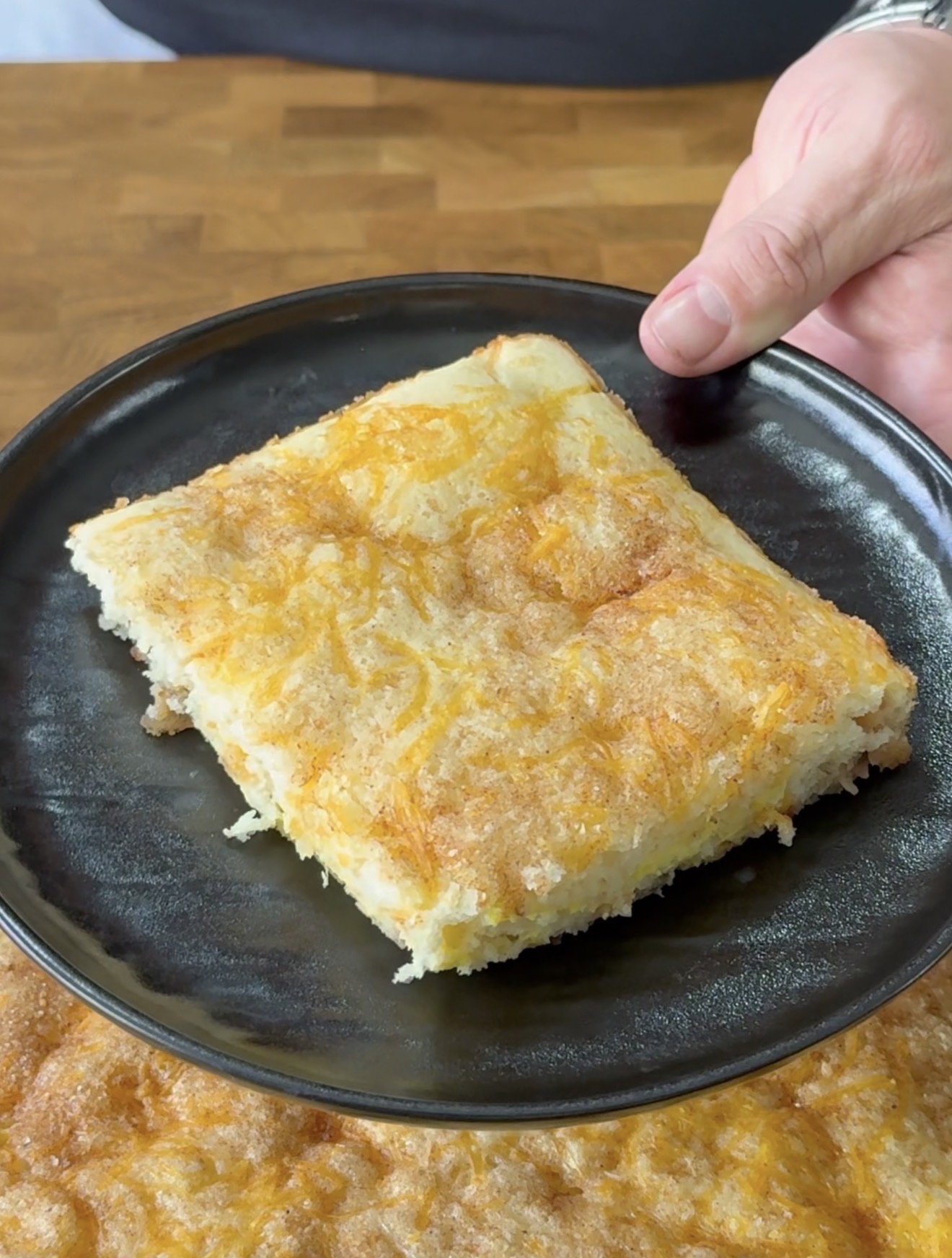 A person holds a black plate with a square slice of cheesy, golden-brown potato casserole. The casserole has a crispy, textured top and moist interior, with more casserole visible on the table below.