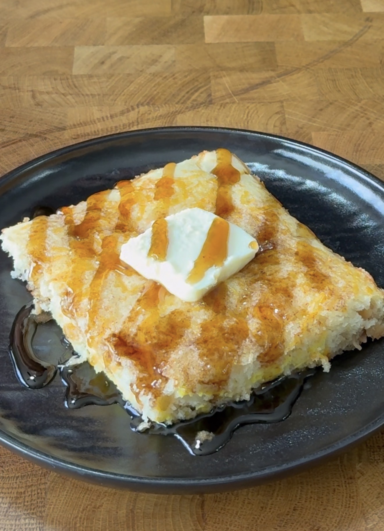 A square piece of baked pancake topped with a pat of butter and drizzled with syrup sits on a dark plate, set on a wooden surface.