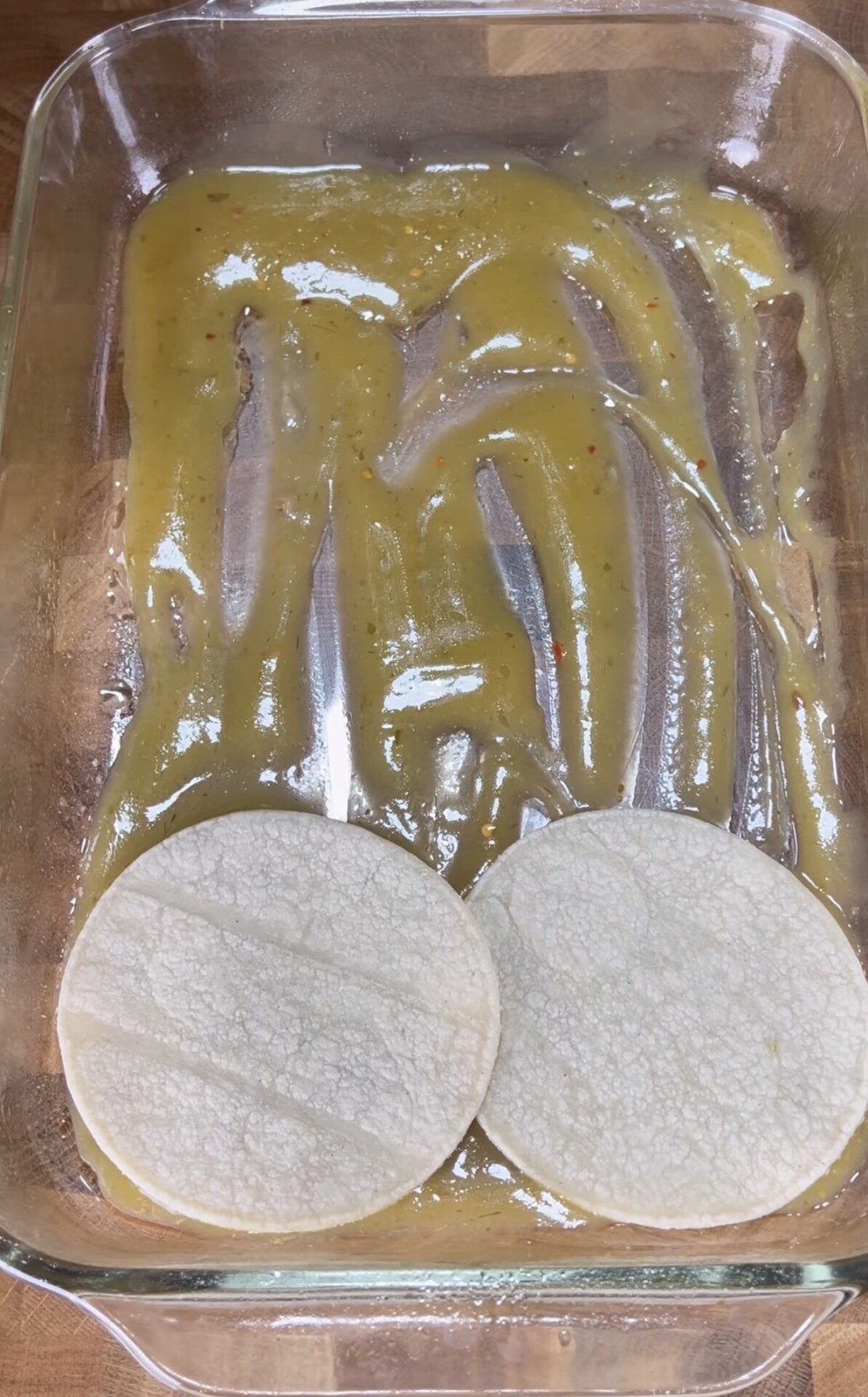 A glass baking dish with green enchilada sauce spread on the bottom and two corn tortillas placed side by side inside the dish.