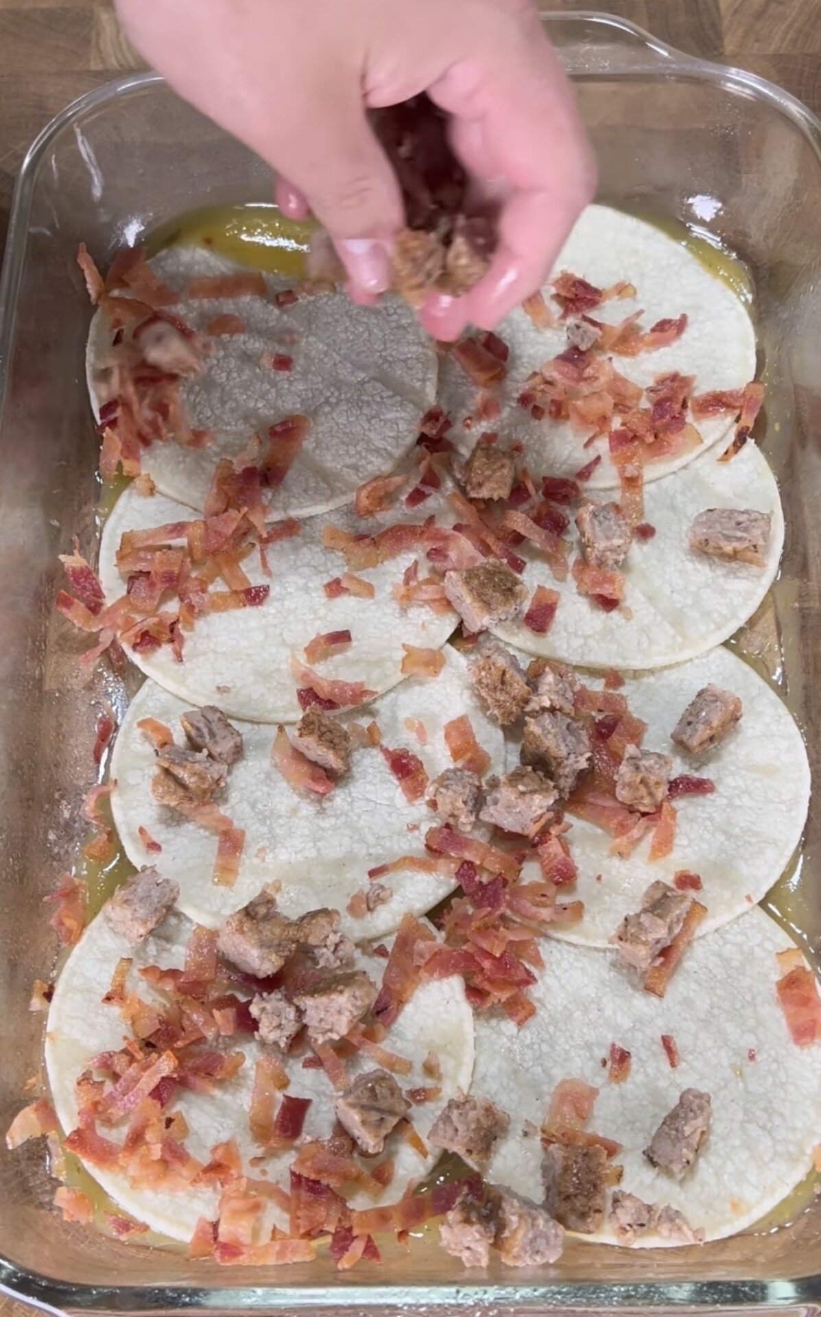 A hand sprinkles sausage pieces over corn tortillas and bacon pieces in a glass baking dish, preparing a layered dish on a wooden surface.