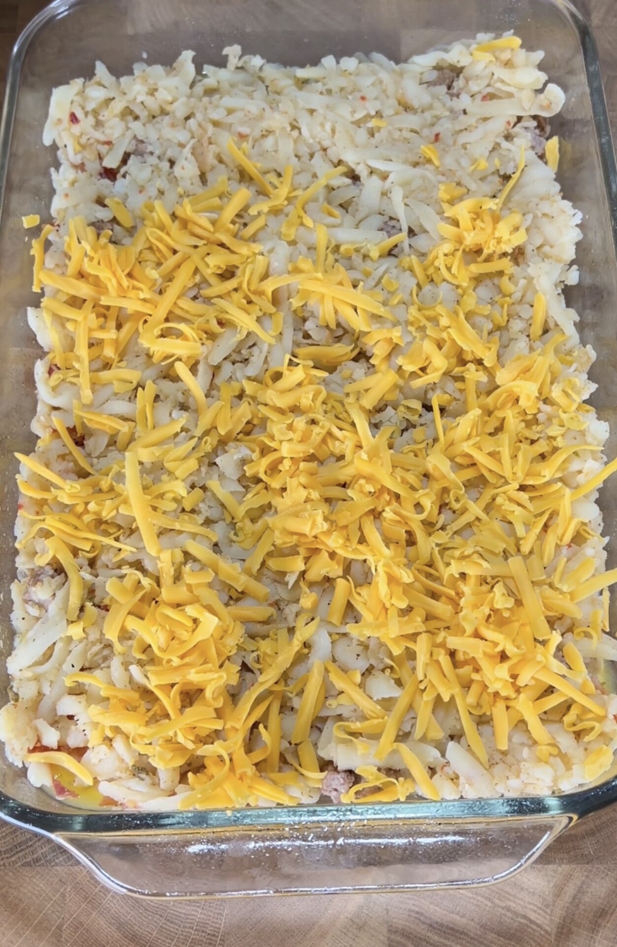 A glass baking dish filled with a mixture of shredded potatoes and ground meat, topped with a layer of shredded yellow cheddar cheese, ready to be baked.