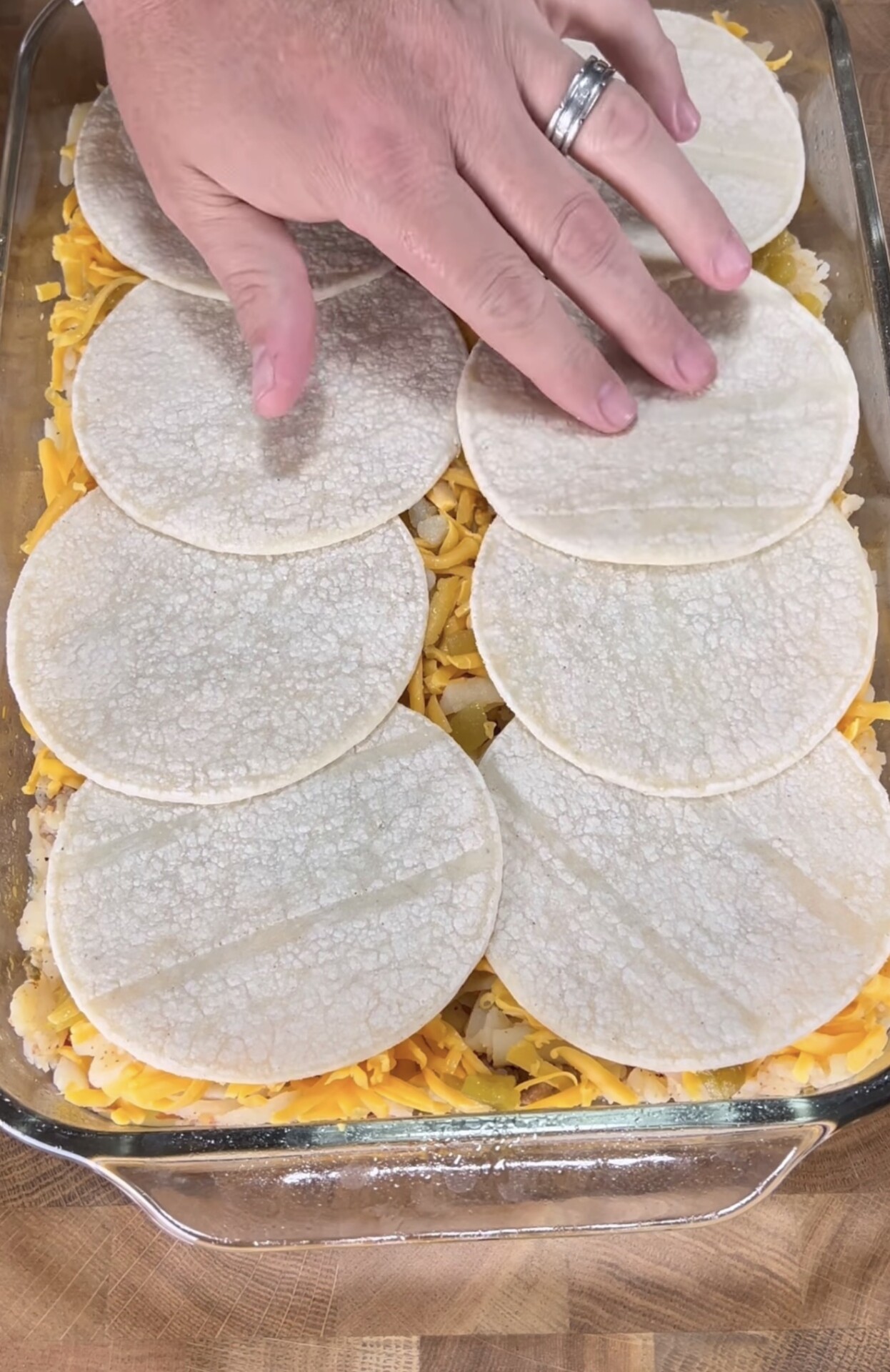 A hand places corn tortillas over a layer of shredded cheese and other ingredients in a glass baking dish, preparing a layered casserole on a wooden surface.