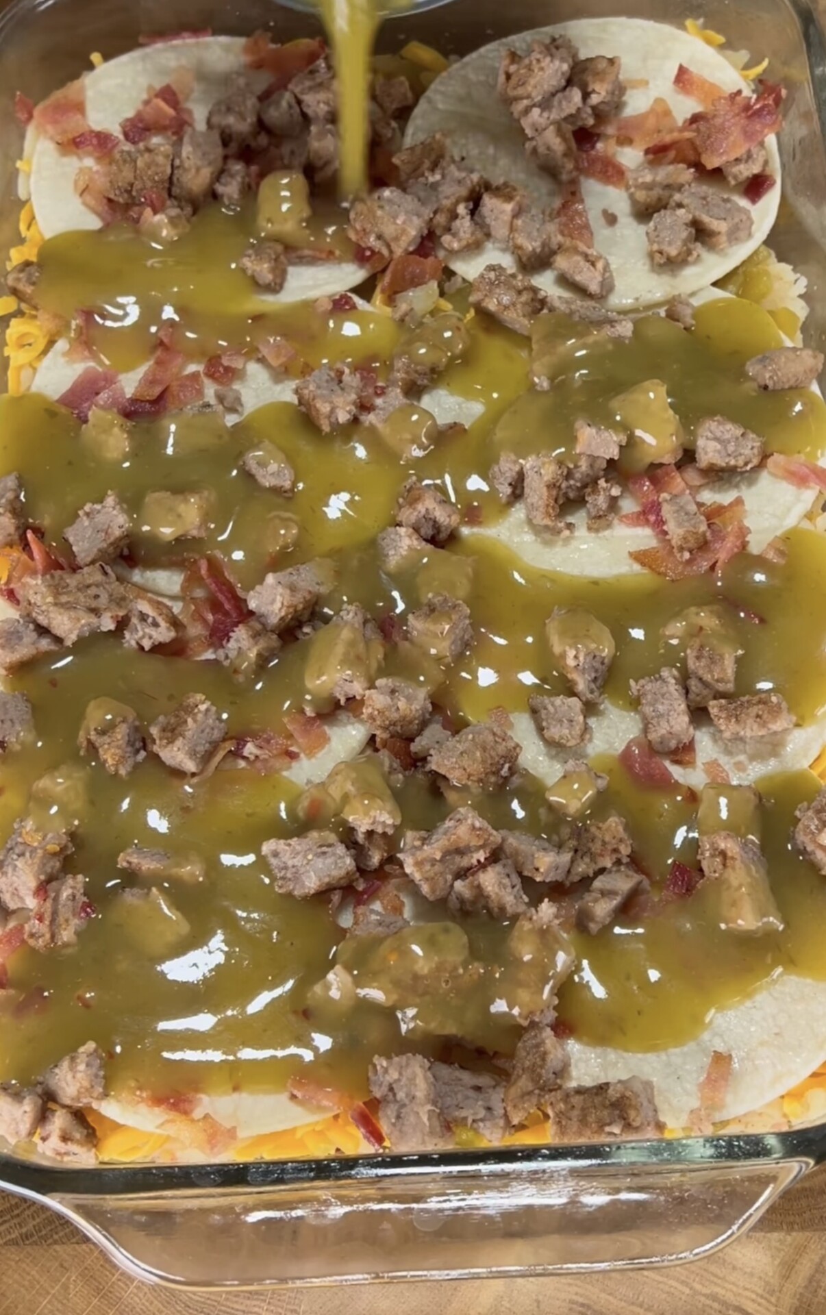 A glass baking dish filled with layered tortillas, shredded cheese, bacon pieces, and sausage chunks, being topped with green enchilada sauce.