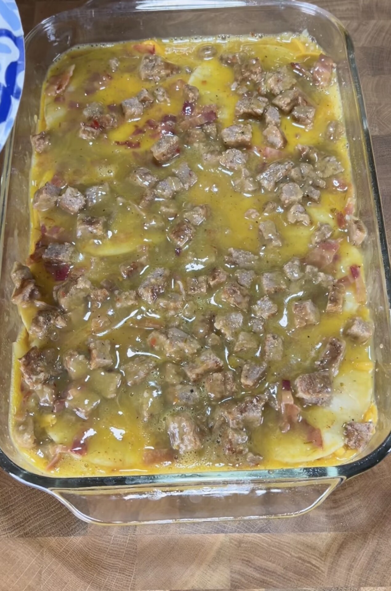 A glass baking dish filled with a casserole topped with chunks of meat and a layer of sauce, resting on a wooden surface. The dish has visible pieces of potatoes and what appears to be melted cheese.