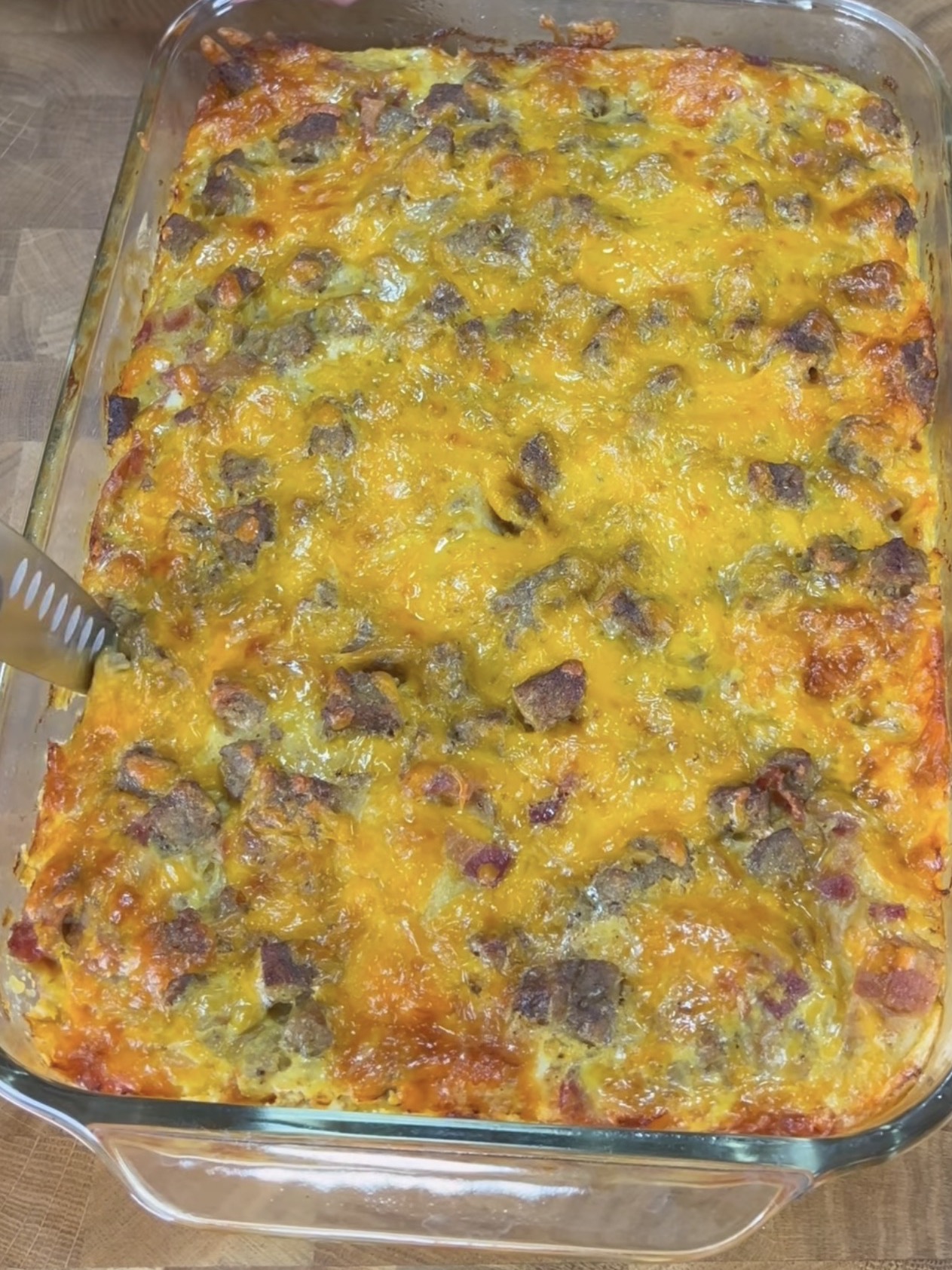 A glass baking dish filled with a freshly baked casserole topped with melted cheddar cheese and pieces of sausage. A serving spatula is visible along the left edge of the dish.