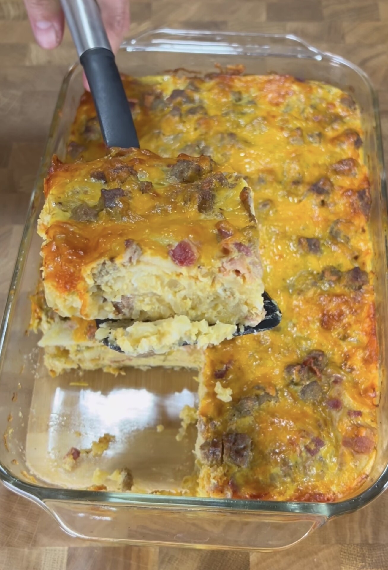 A hand lifts a square serving of cheesy breakfast casserole from a glass baking dish filled with more casserole, showing layers of eggs, cheese, and sausage on a wooden surface.