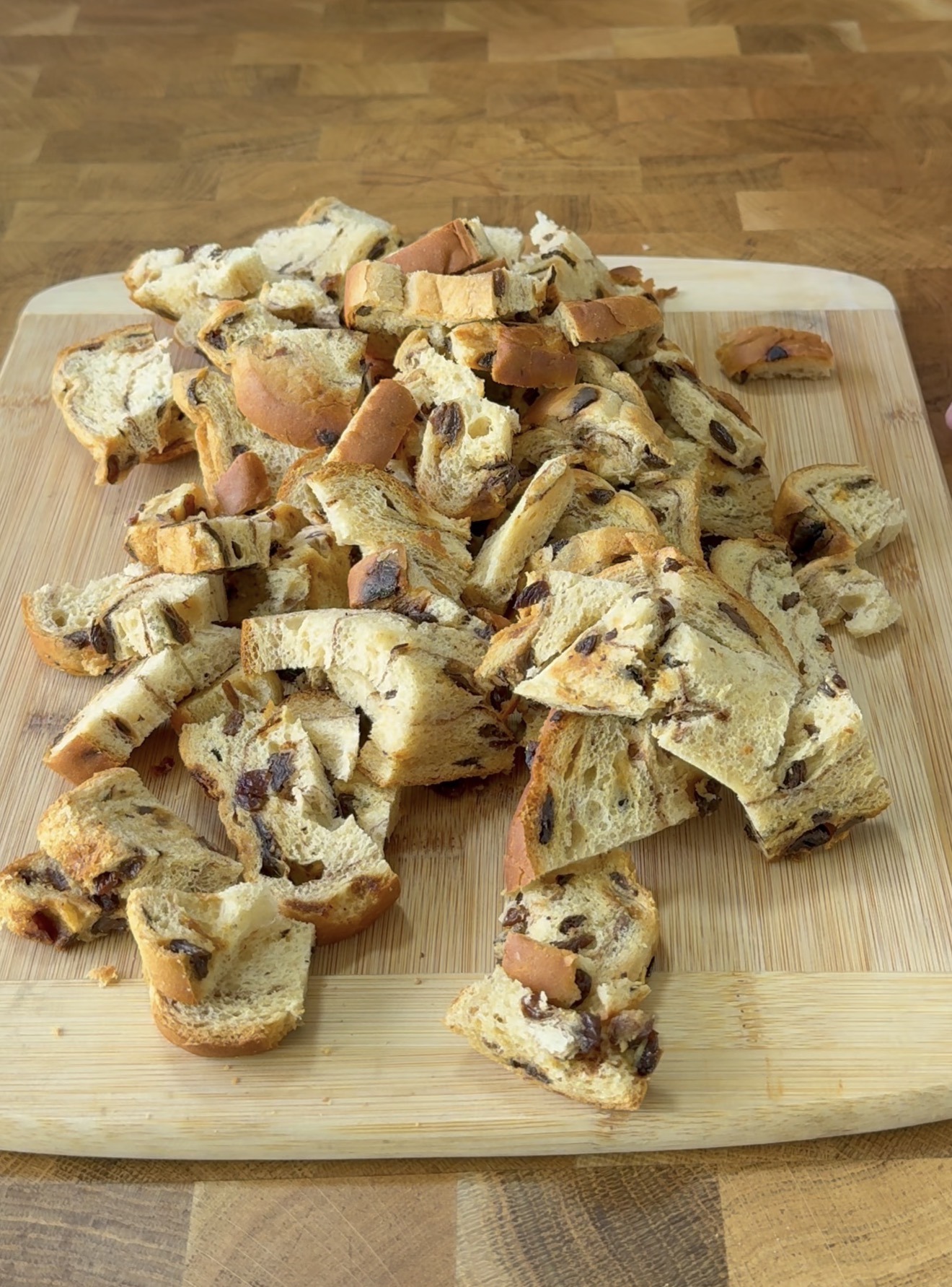 Chunks of sliced raisin bread are scattered on a wooden cutting board, placed on a wooden countertop.
