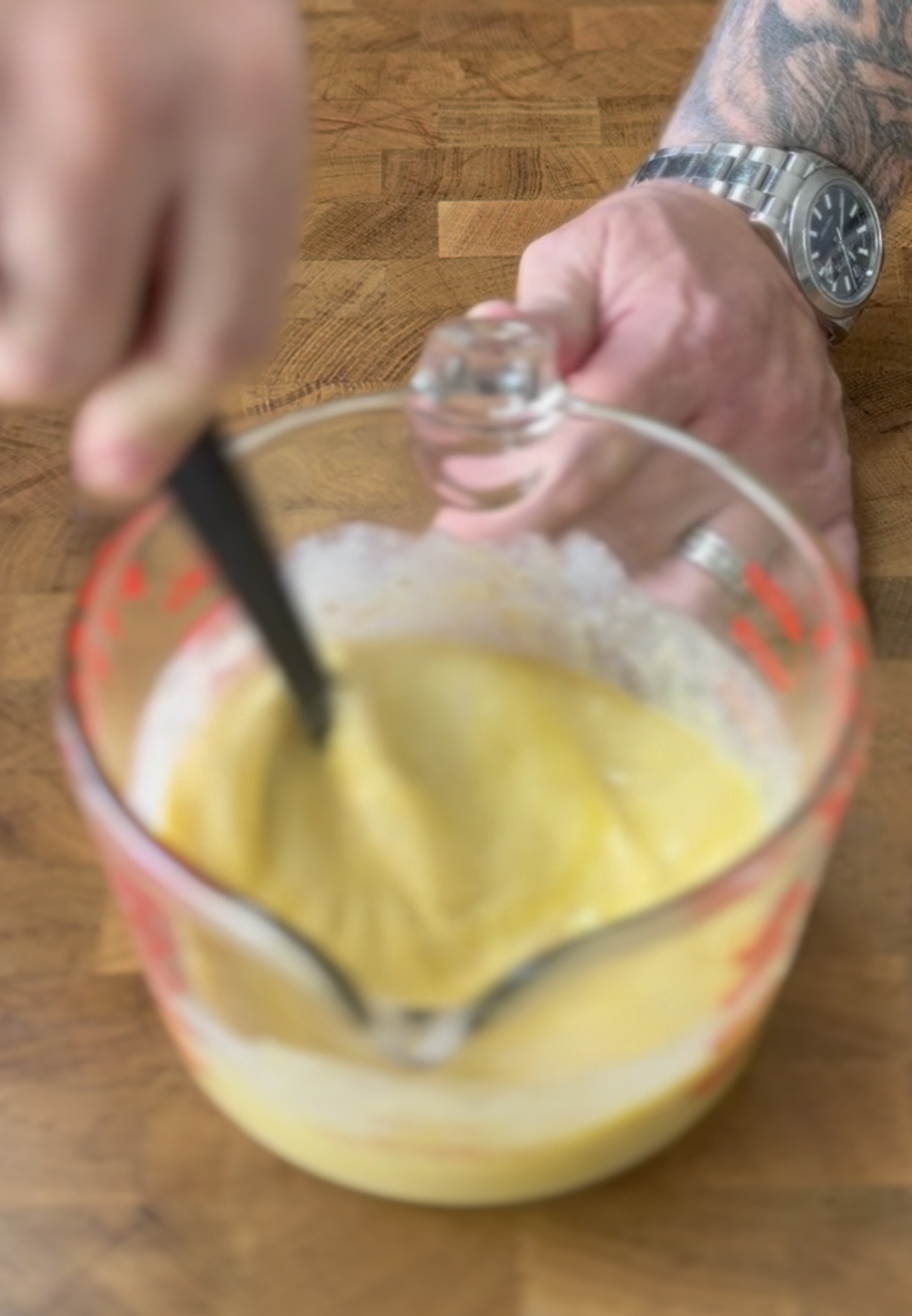 A person with a watch and tattoo on their arm is whisking a yellow batter in a glass measuring cup on a wooden surface.