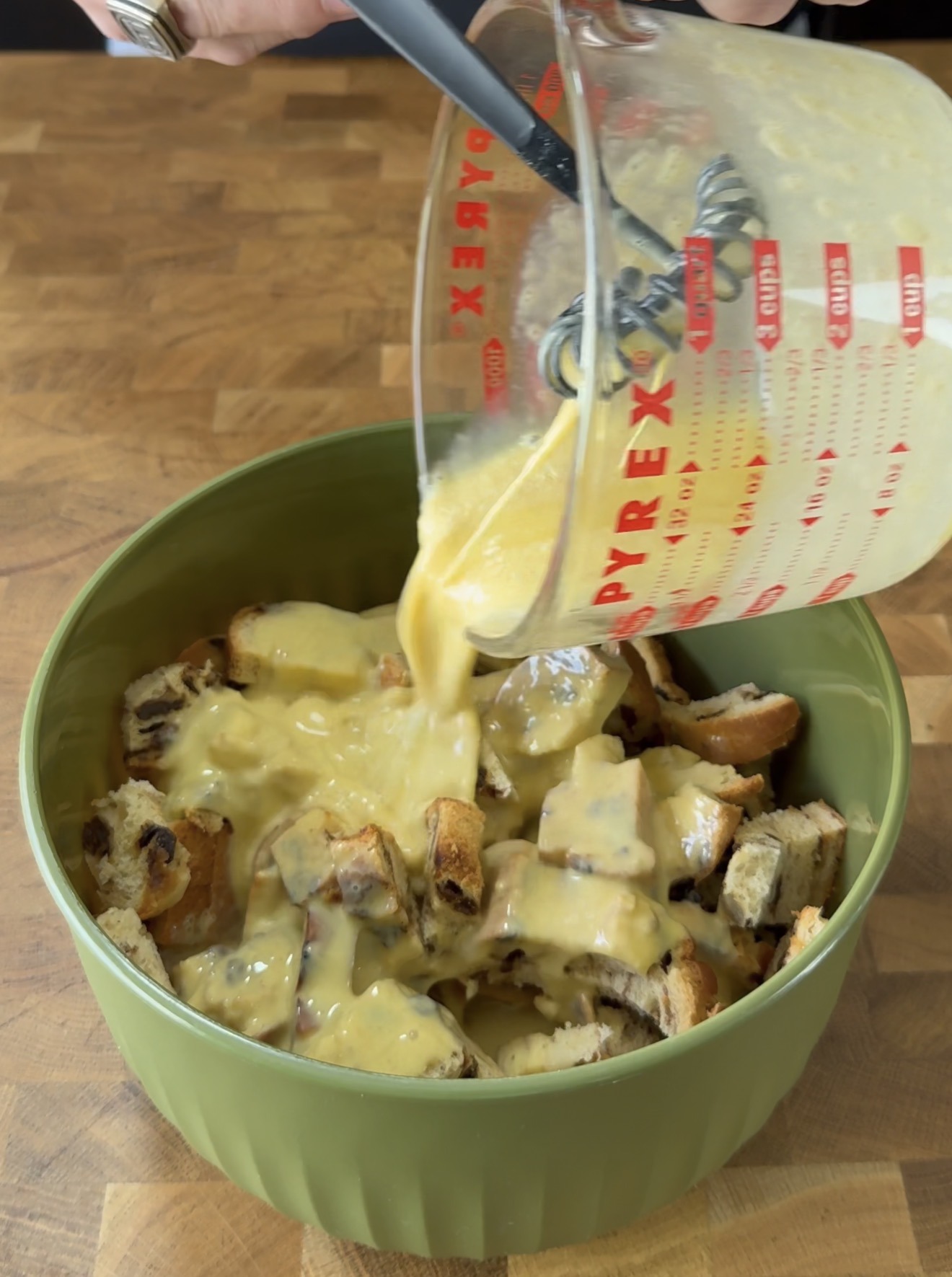 A hand pours a yellow egg mixture from a glass measuring cup over cubed bread in a green mixing bowl on a wooden surface.