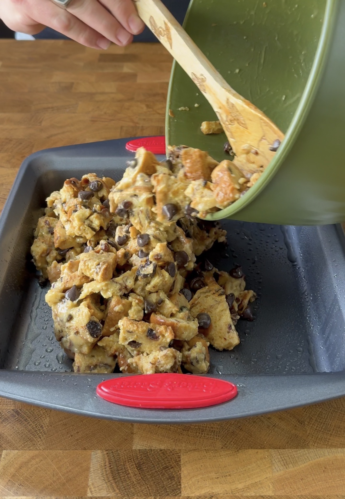 A person pours a bowl of bread pudding mixture with chocolate chips into a greased baking pan on a wooden countertop.