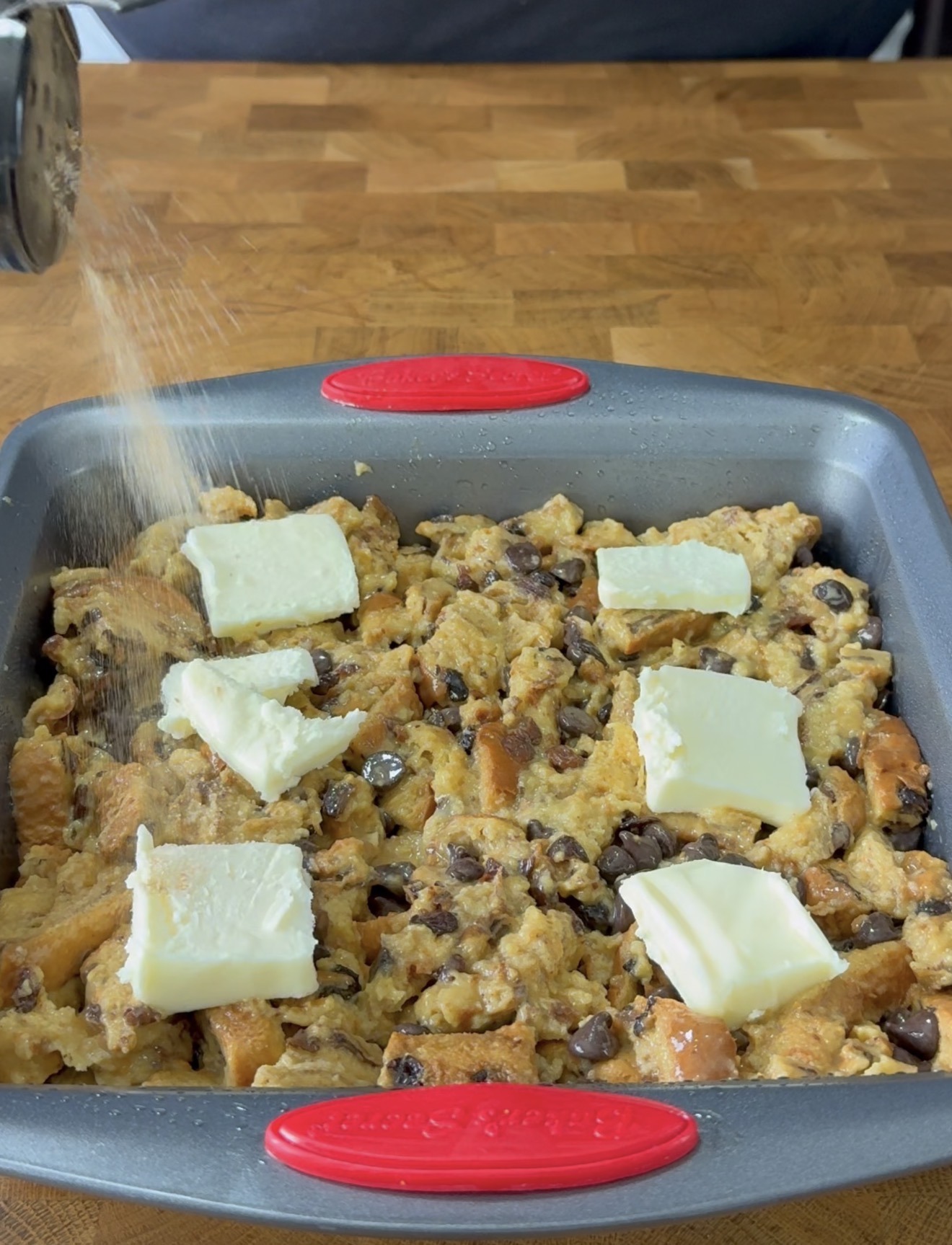 A square baking pan filled with unbaked bread pudding mixture, topped with chunks of butter. Sugar is being sprinkled over the top. The pan sits on a wooden surface.