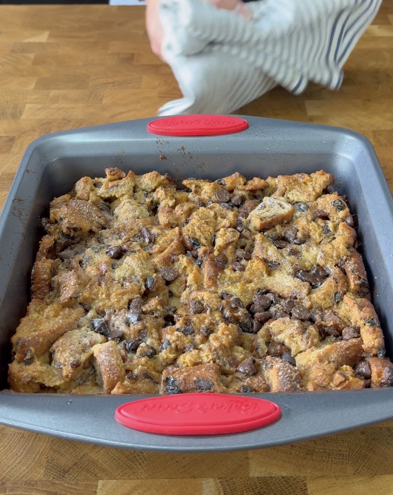 A baked bread pudding with chocolate chips sits in a square gray baking pan with red handles on a wooden surface. A hand holding a striped cloth appears in the background.