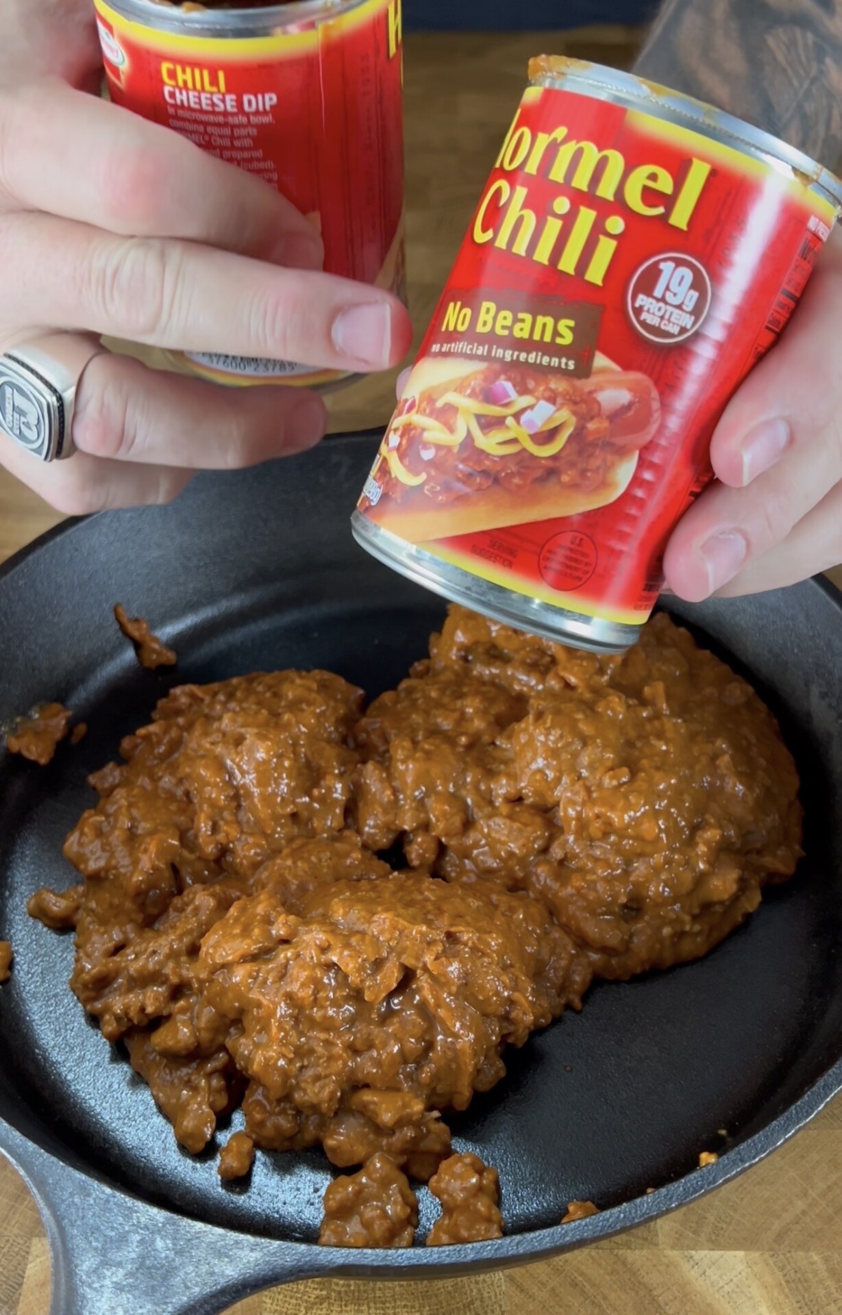 A person holds cans of Hormel Chili and chili cheese dip above a skillet filled with heated, chunky chili on a wooden surface.