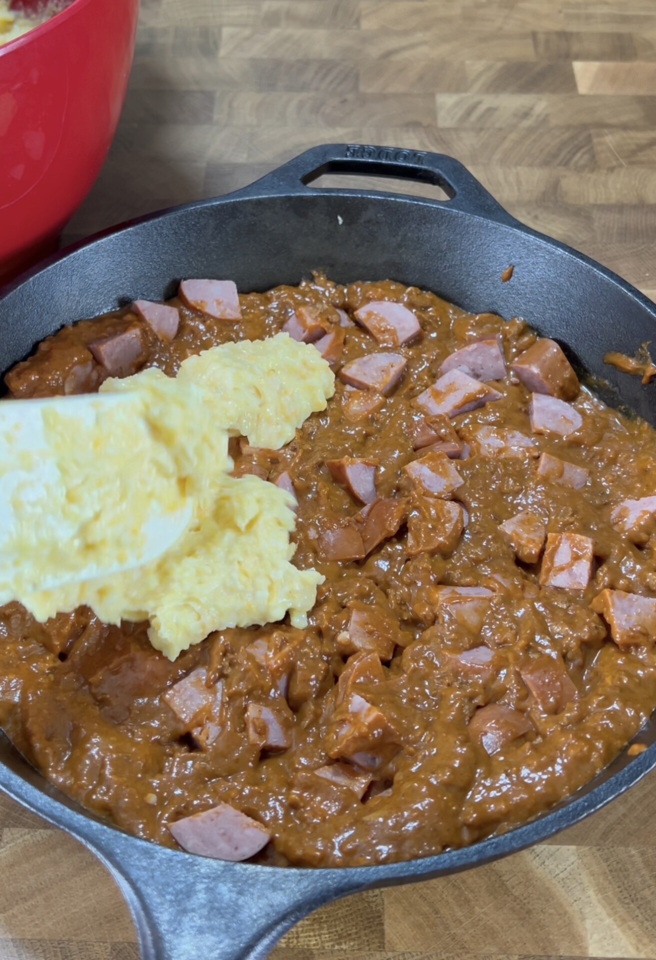 A cast iron skillet filled with a cooked mixture of diced sausage and a thick, reddish-brown sauce sits on a wooden surface. Scrambled eggs are being added to the skillet from the left side.