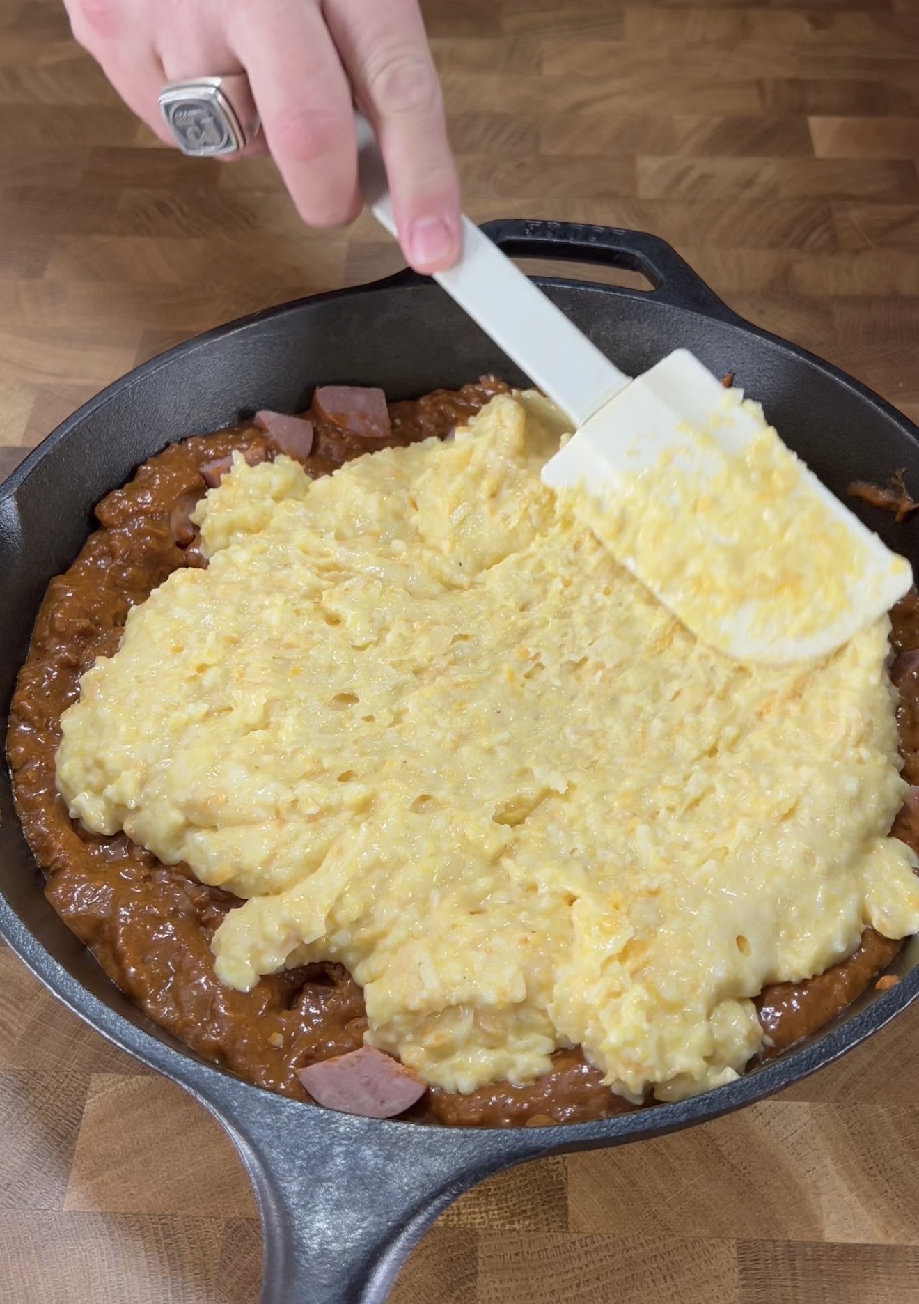 A hand holds a white spatula, spreading creamy scrambled eggs over baked beans and pieces of sausage in a black cast iron skillet on a wooden surface.