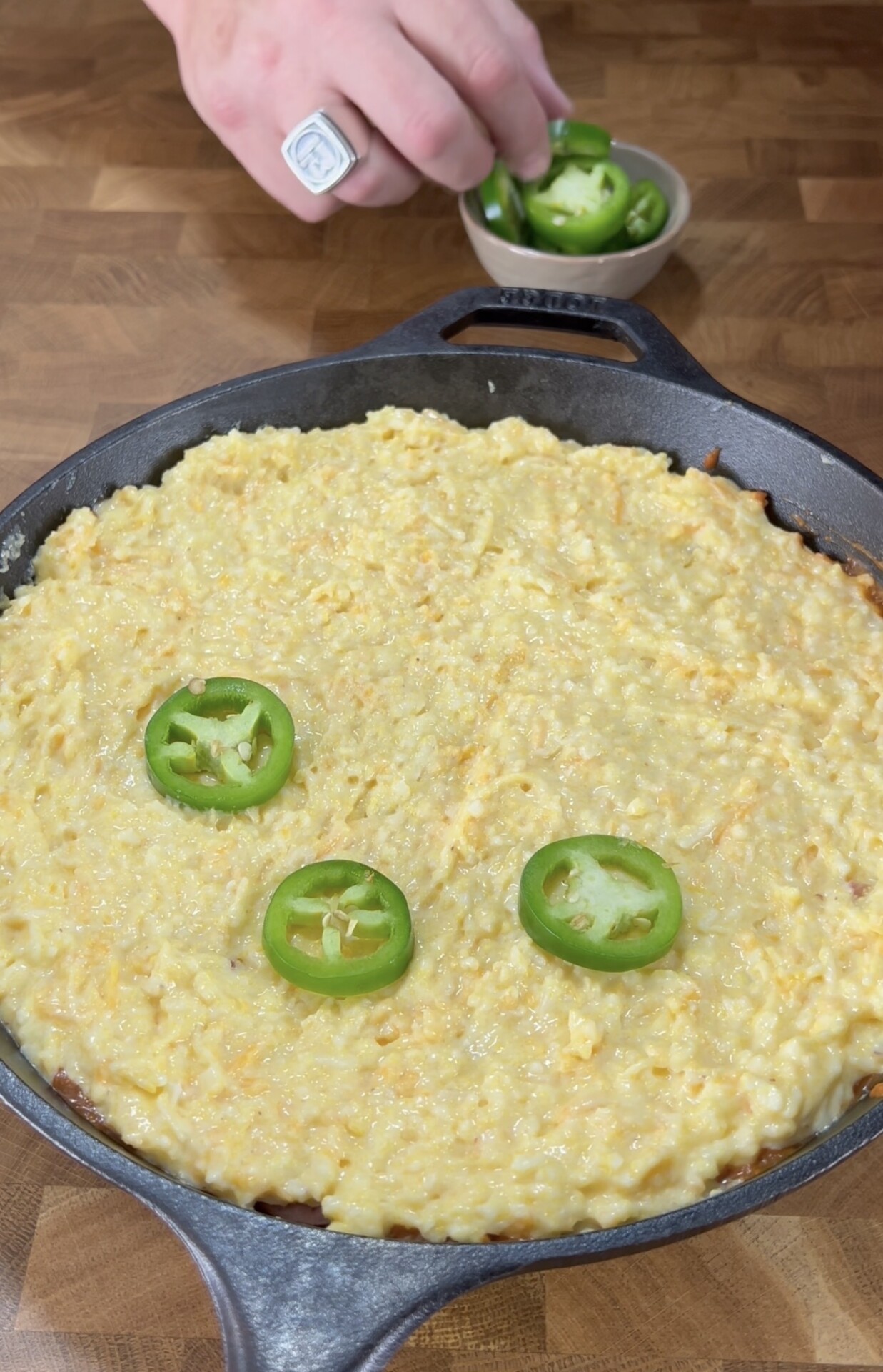A hand holding a bowl of sliced jalapeños hovers over a cast iron skillet filled with cheesy cornbread batter topped with three jalapeño slices on a wooden countertop.