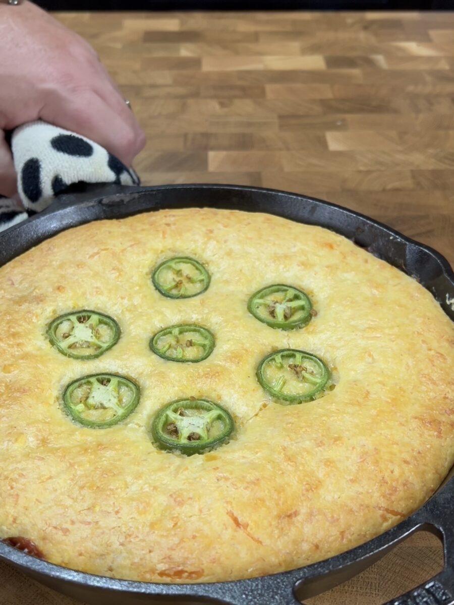 A golden, baked cornbread topped with sliced jalapeños in a cast iron skillet sits on a wooden surface. A hand with a towel holds the skillet handle.