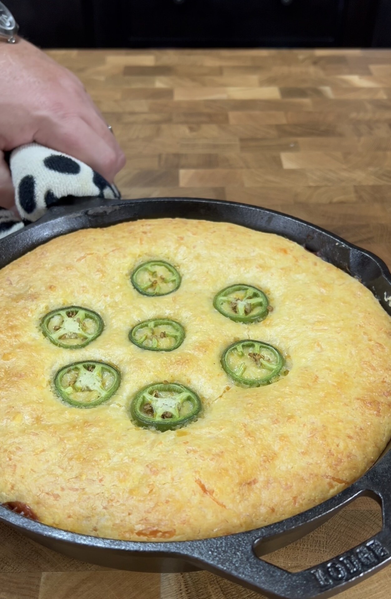 A person holds a skillet with a golden baked cornbread topped with sliced jalapeños, resting on a wooden countertop.