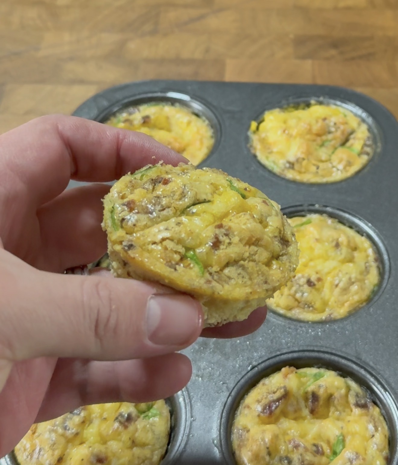 A hand holds a baked egg muffin with visible sausage and vegetables above a muffin tin filled with similar egg muffins on a wooden surface.