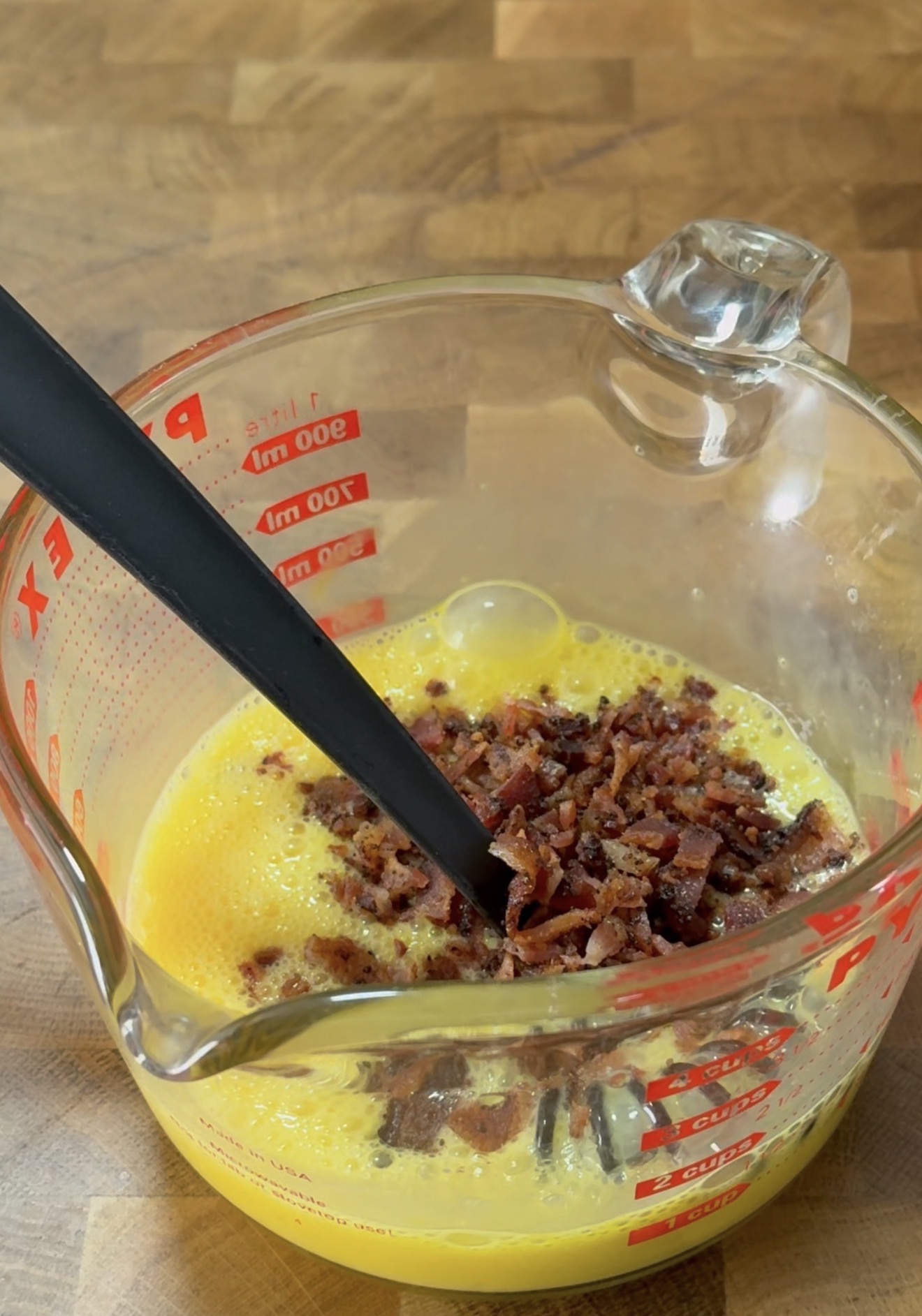 A glass measuring cup with beaten eggs and crumbled bacon inside sits on a wooden surface. A black spatula rests in the mixture, ready for stirring.