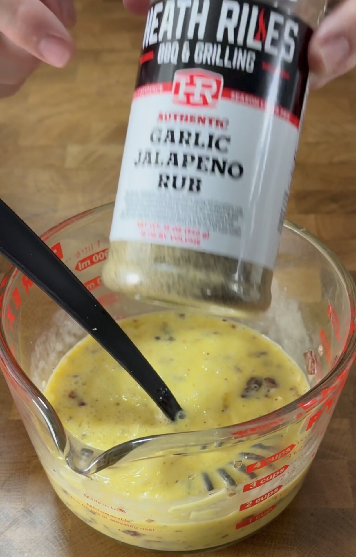 A hand holds a bottle of Heath Riles Garlic Jalapeno Rub above a glass measuring cup containing a yellow egg mixture with a black spoon inside.