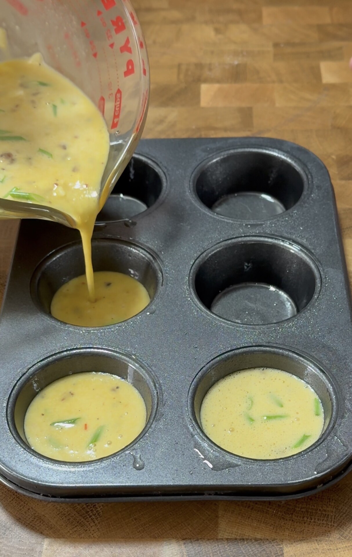 A close-up of a muffin pan with some cups filled as egg mixture with vegetables is being poured from a measuring cup into one of the cups on a wooden countertop.