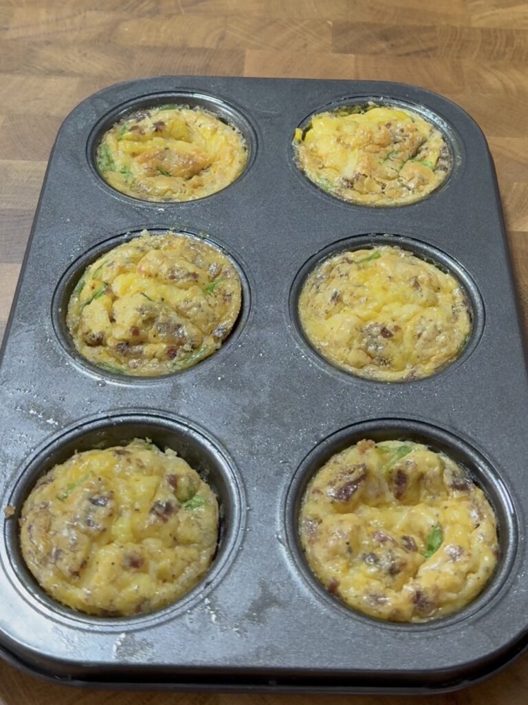 A muffin tin with six baked egg muffins, filled with ingredients like cheese, sausage, and green vegetables, sits on a wooden surface.