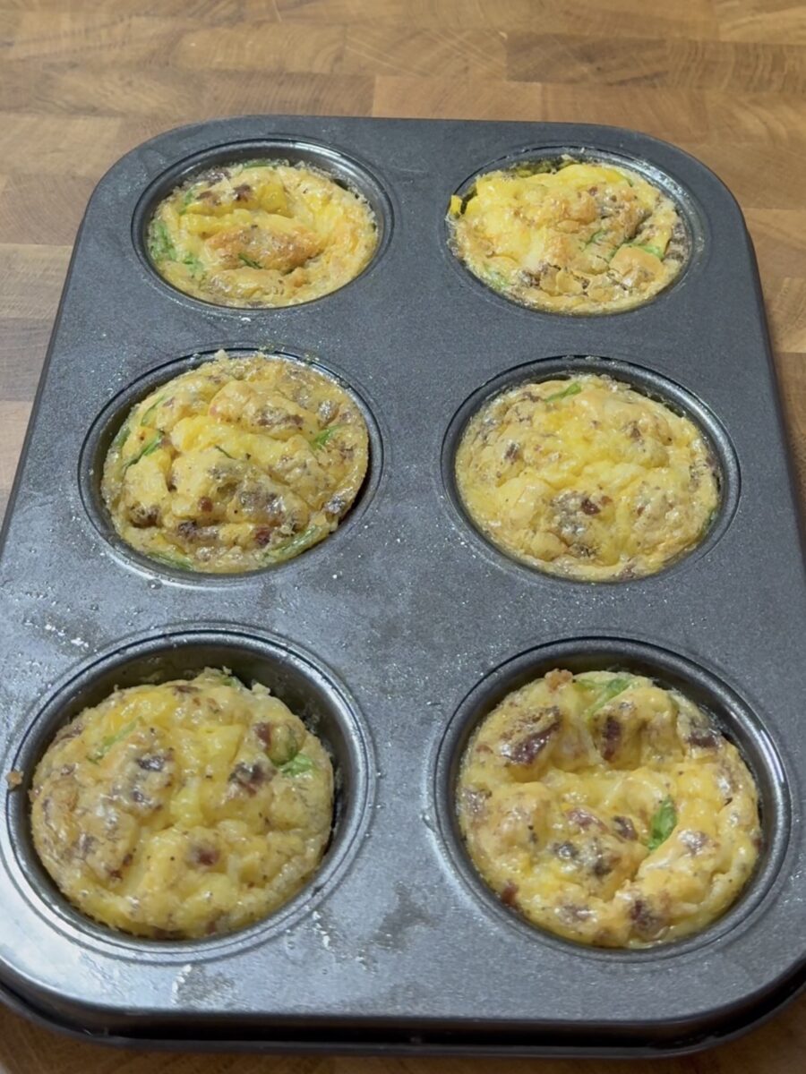 A muffin tin with six baked egg muffins, filled with ingredients like cheese, sausage, and green vegetables, sits on a wooden surface.