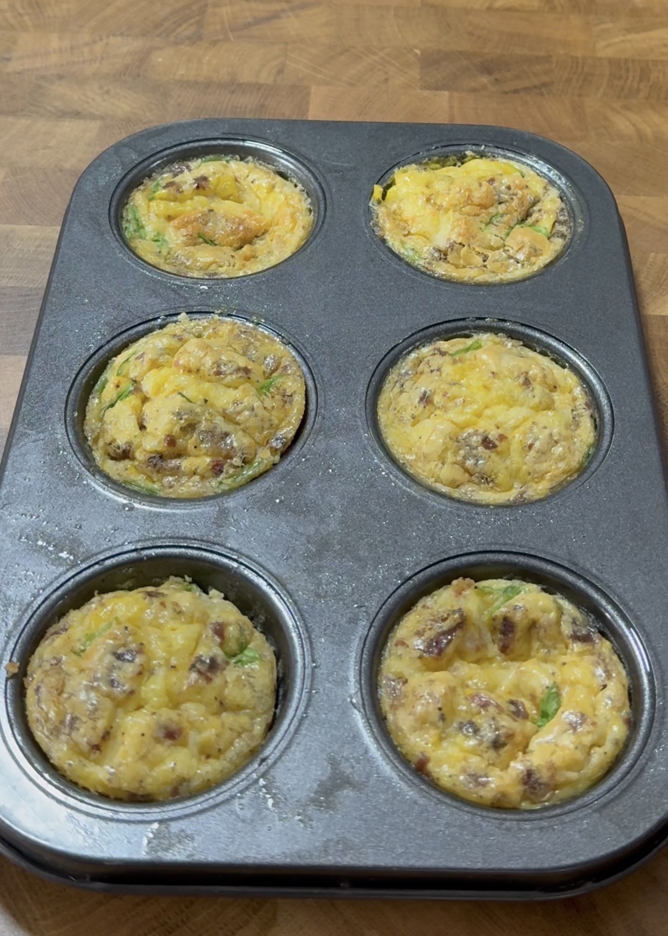 A muffin tin filled with six baked egg muffins containing visible pieces of vegetables, cheese, and sausage, resting on a wooden countertop.