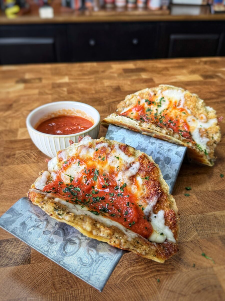 Two taco-shaped dishes made with a baked cheese shell, filled with tomato sauce and melted cheese, are displayed on a wooden counter. A small bowl of marinara sauce sits nearby for dipping.